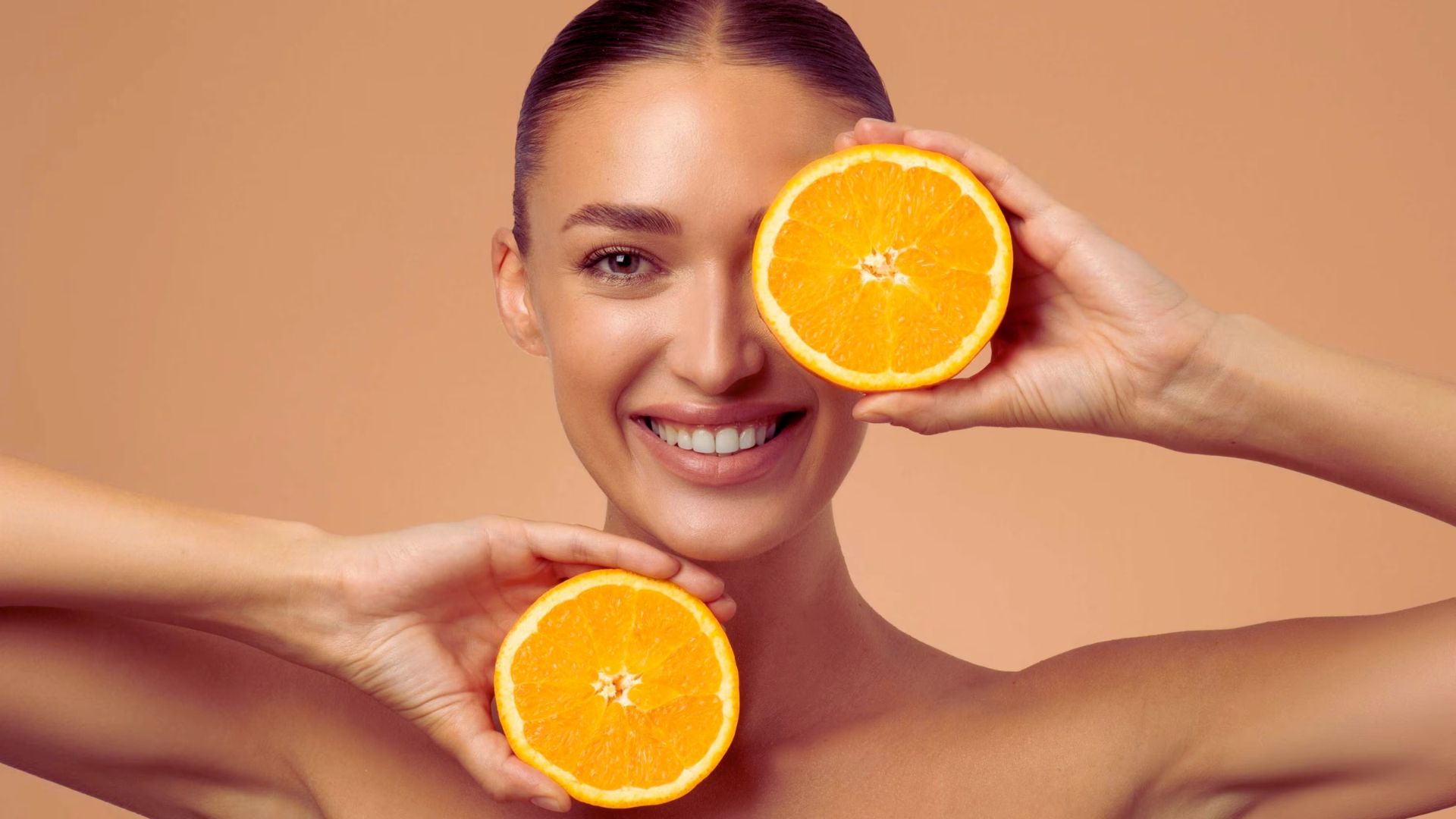 A smiling woman holds two orange halves, one near her face and the other below her chin, against a peach background. The image conveys freshness and joy.