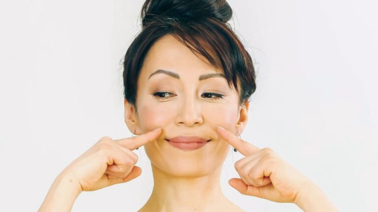 Woman with hair in a bun gently presses cheeks with fingers, smiling slightly. She wears a neutral expression against a plain white background, suggesting focus.