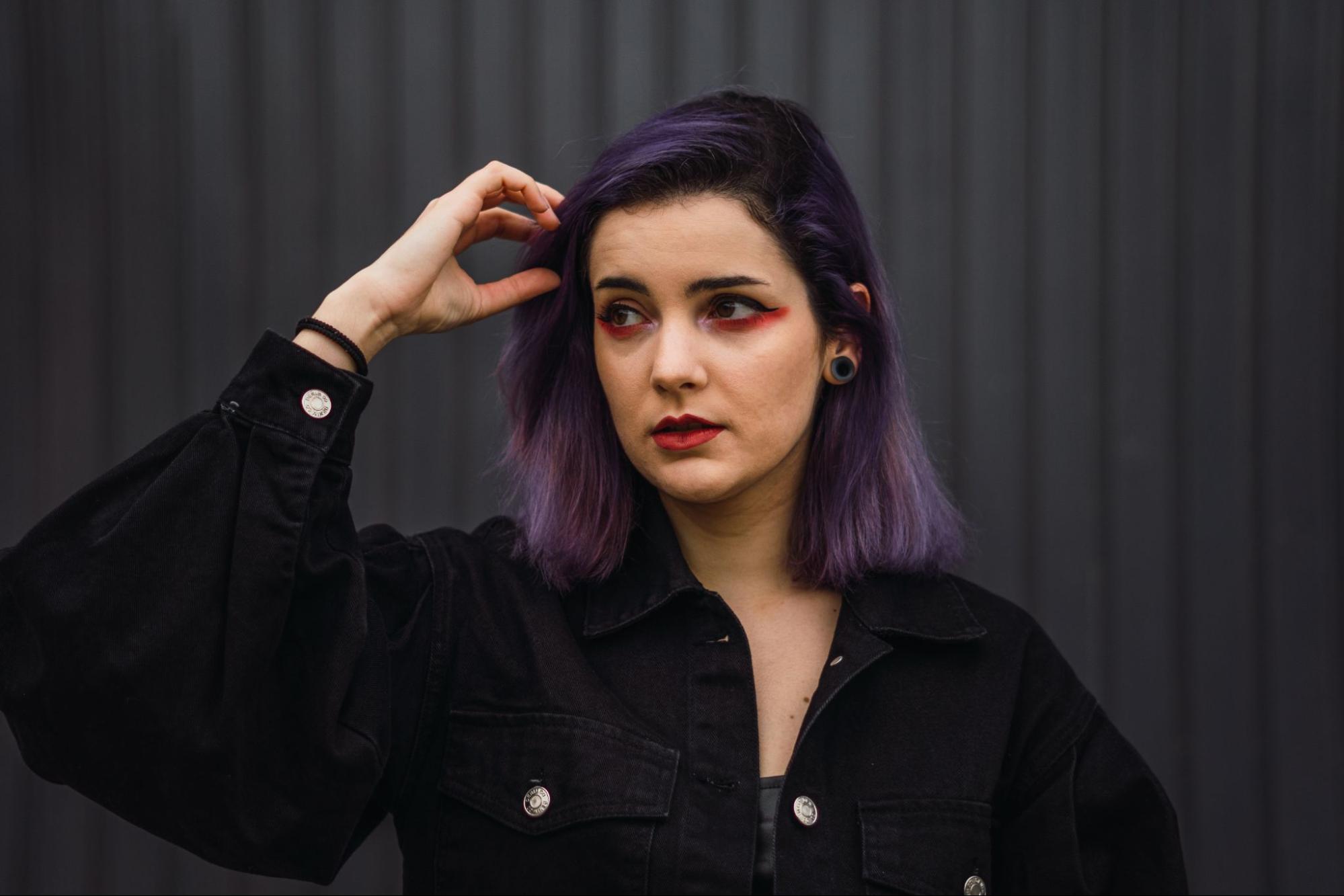 A woman with striking purple hair dressed in a sleek black jacket poses with a confident expression.
