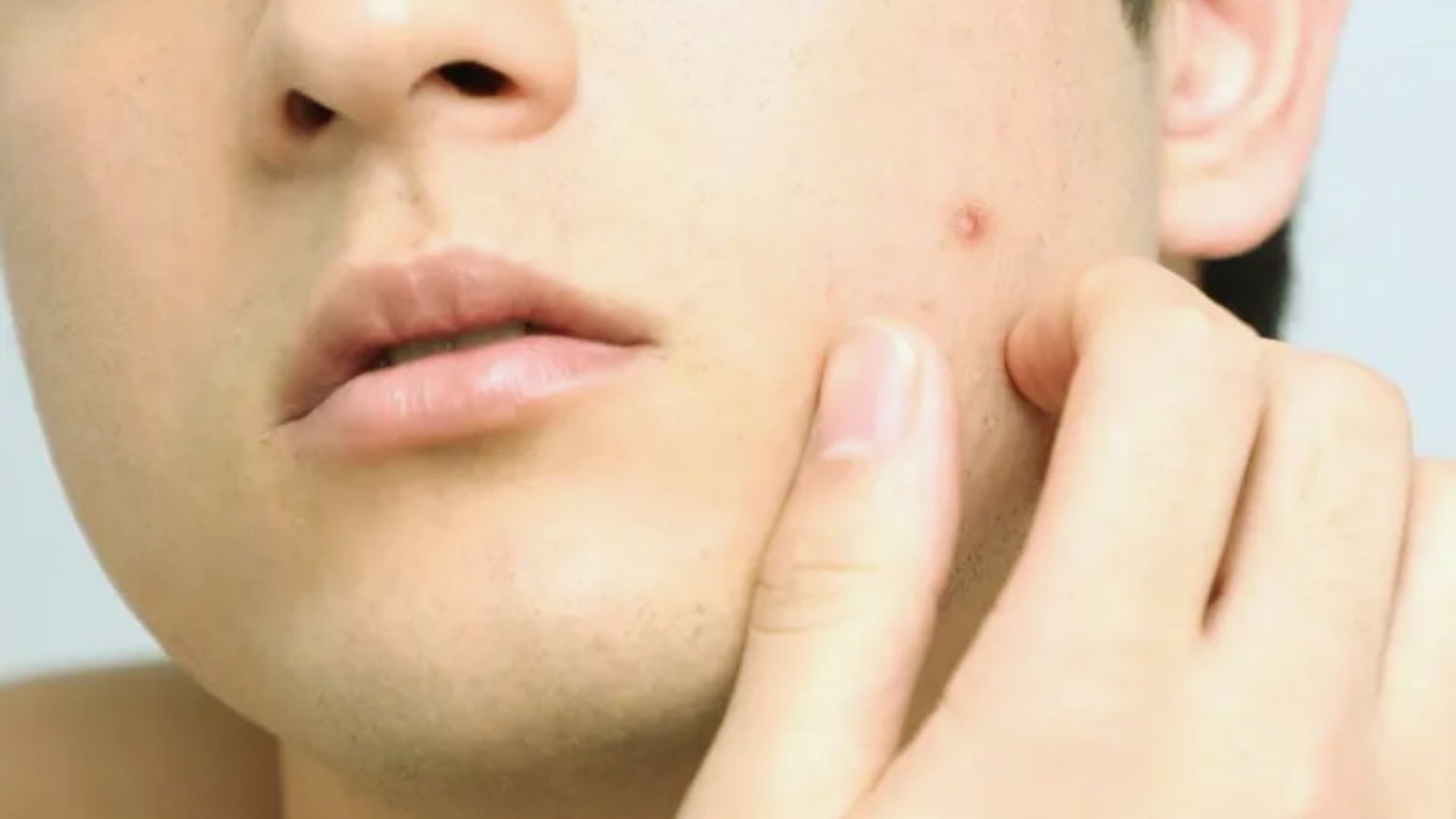 Close-up of a person touching a small pimple on their cheek. The skin is smooth, highlighting the blemish. The mood is neutral and focused.