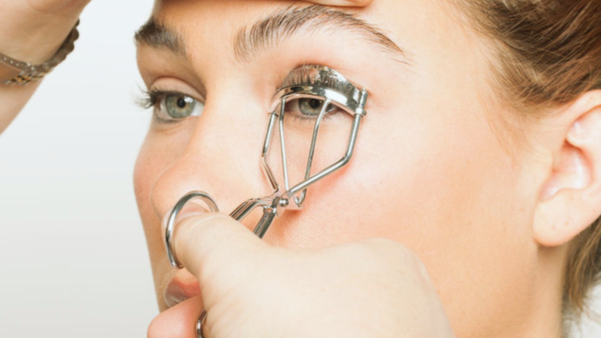 A person uses an eyelash curler on their right eye. The background is soft and neutral, focusing on the eye and curler. The scene conveys concentration.
