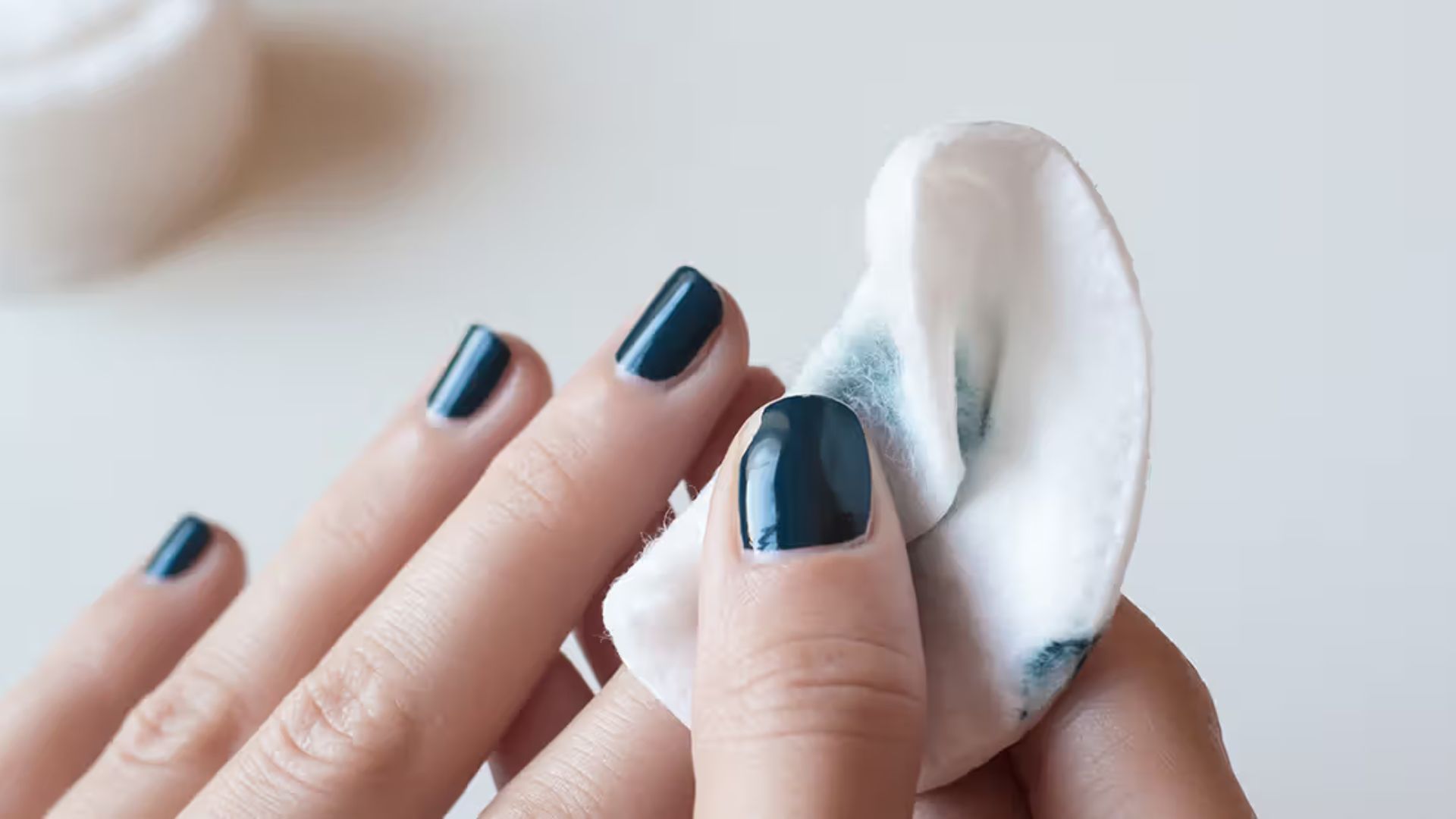 Close-up of hands removing dark nail polish with a cotton pad. The focus is on clean fingernails on one hand and the removal process on the other.