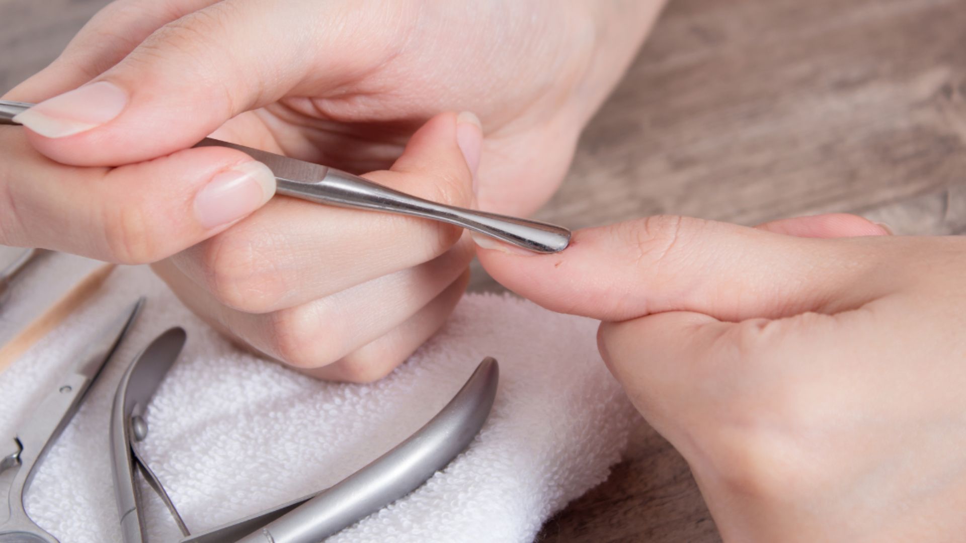 Close-up of a hand receiving a manicure. Another hand uses a metal tool to gently push back cuticles. Manicure tools rest on a towel, creating a calm and clean atmosphere.