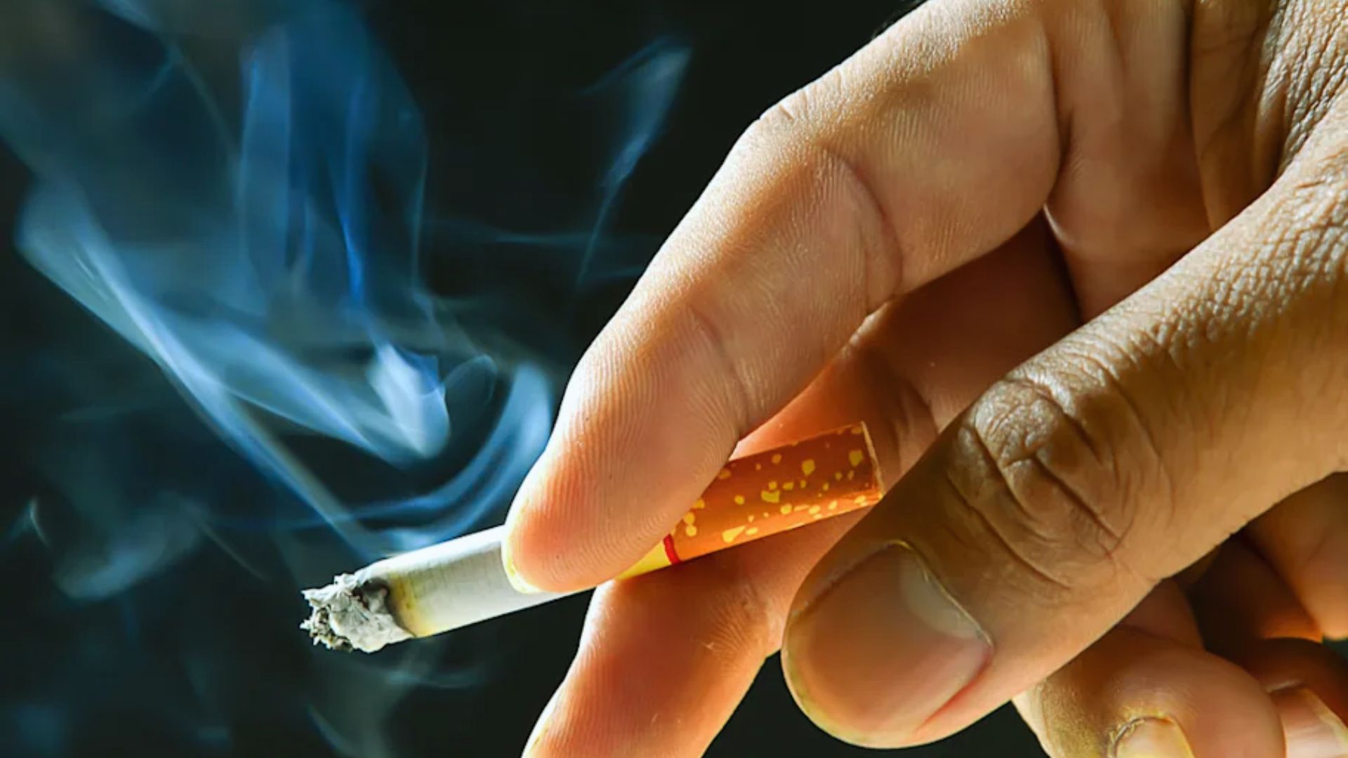 A close-up of a hand holding a lit cigarette with smoke curling upwards against a black background, creating a tense and moody atmosphere.