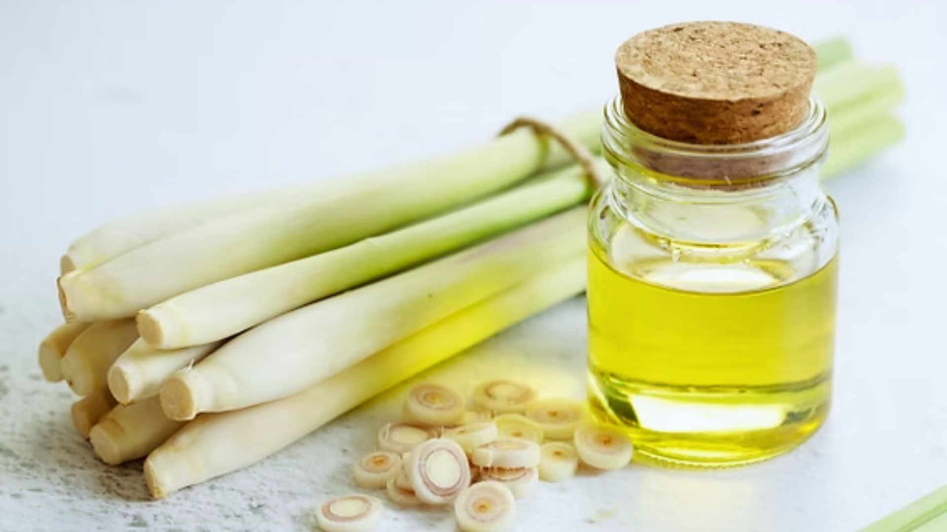 Fresh lemongrass stalks lie beside a small glass jar filled with yellow oil, corked, with sliced lemongrass scattered nearby on a white surface.