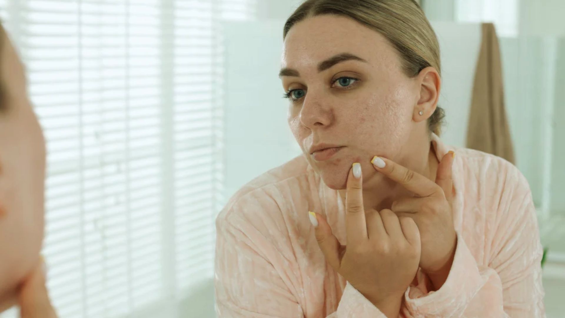 A woman in a pink robe examines her face closely in the mirror, gently touching her cheek. The setting is a well-lit bathroom with blinds and a towel in the background.