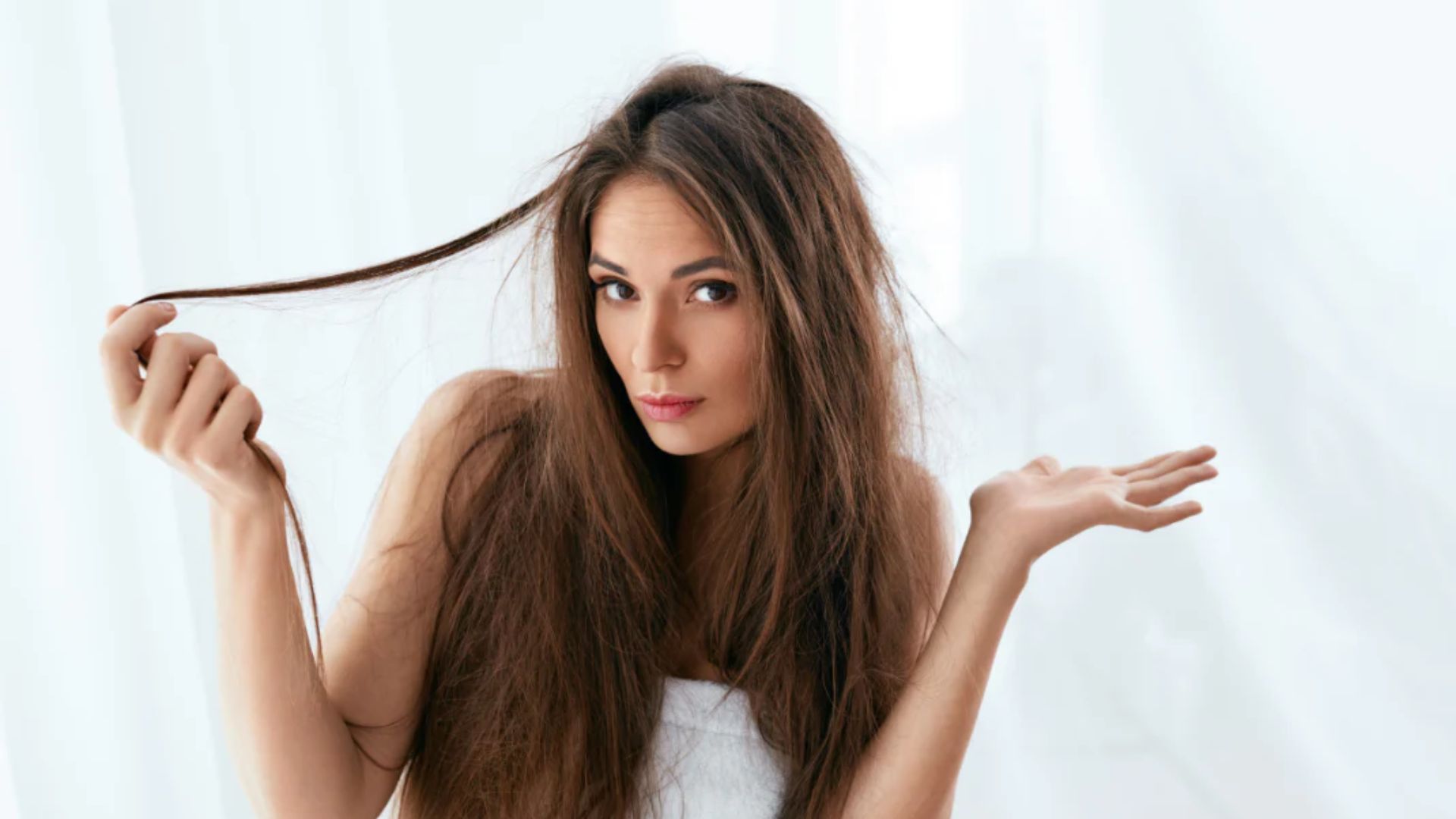 A woman with long, tousled brown hair looks puzzled, holding a strand of hair. She wears a white top against a soft white background, suggesting confusion.