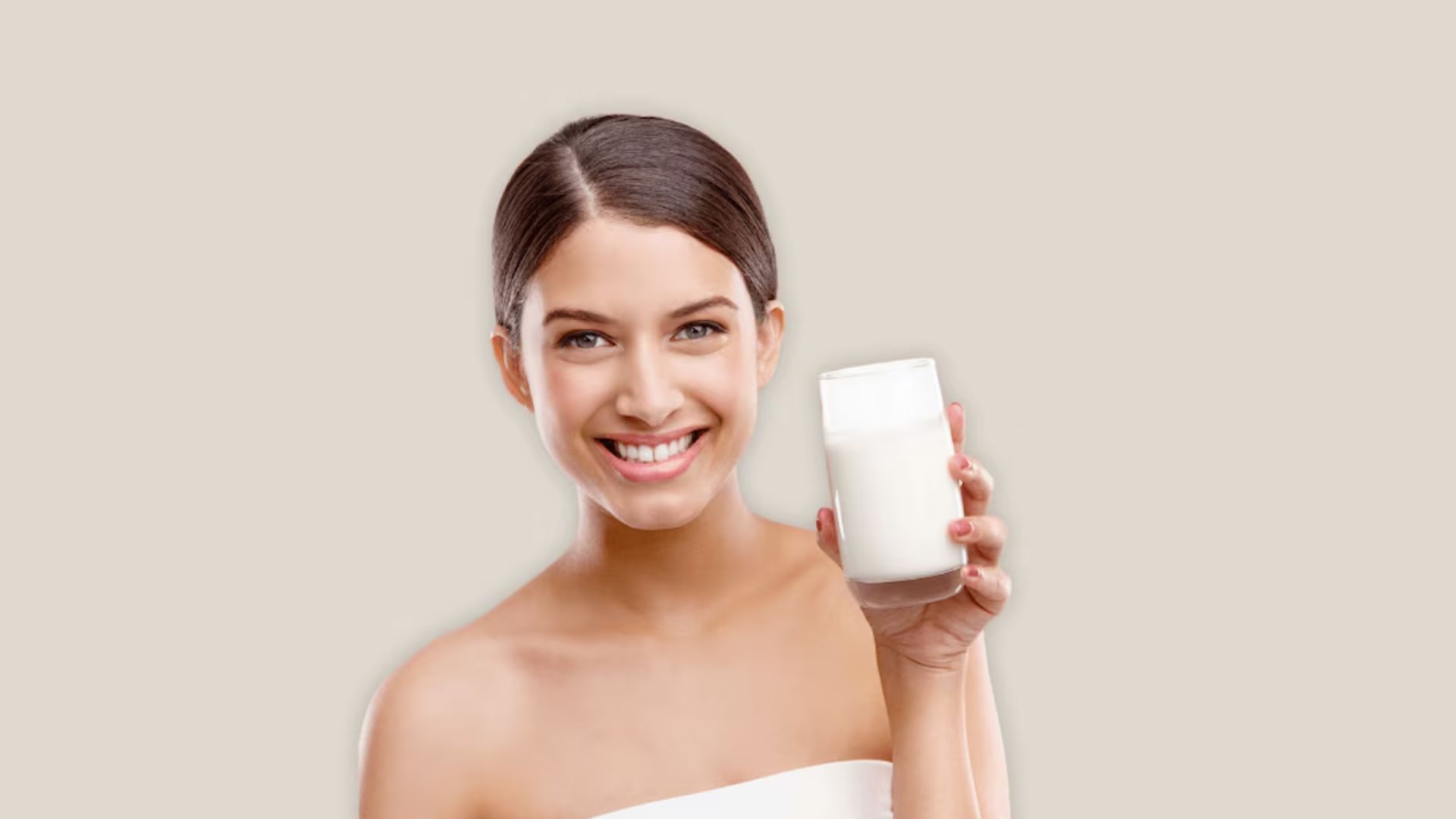 Smiling woman holding a glass of milk, wearing a strapless top. The background is plain beige, conveying a fresh and inviting tone.