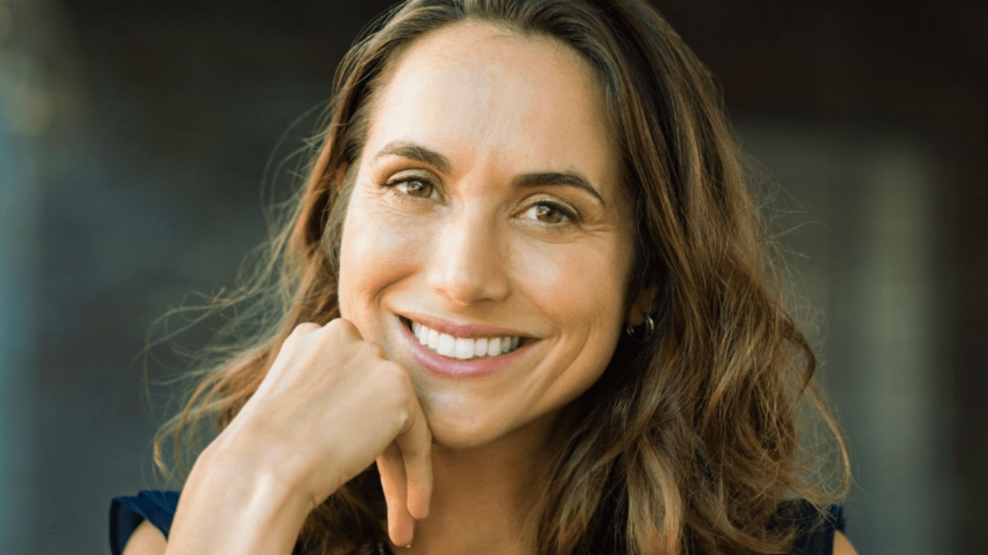 Smiling woman with long, wavy brown hair, resting her chin on her hand. She exudes warmth and friendliness against a blurred, dark background.