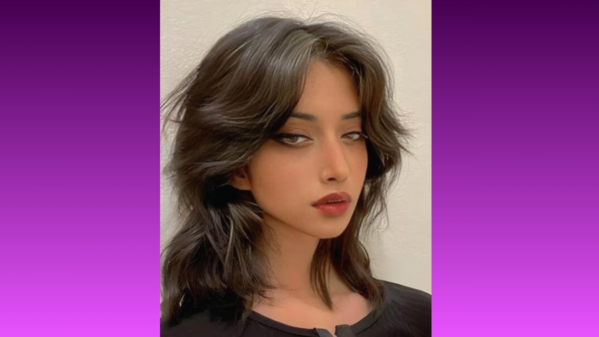 Portrait of a person with layered, wavy black hair and dramatic makeup featuring bold eyeliner and dark red lips, set against a purple gradient background. The expression is confident and stylish.