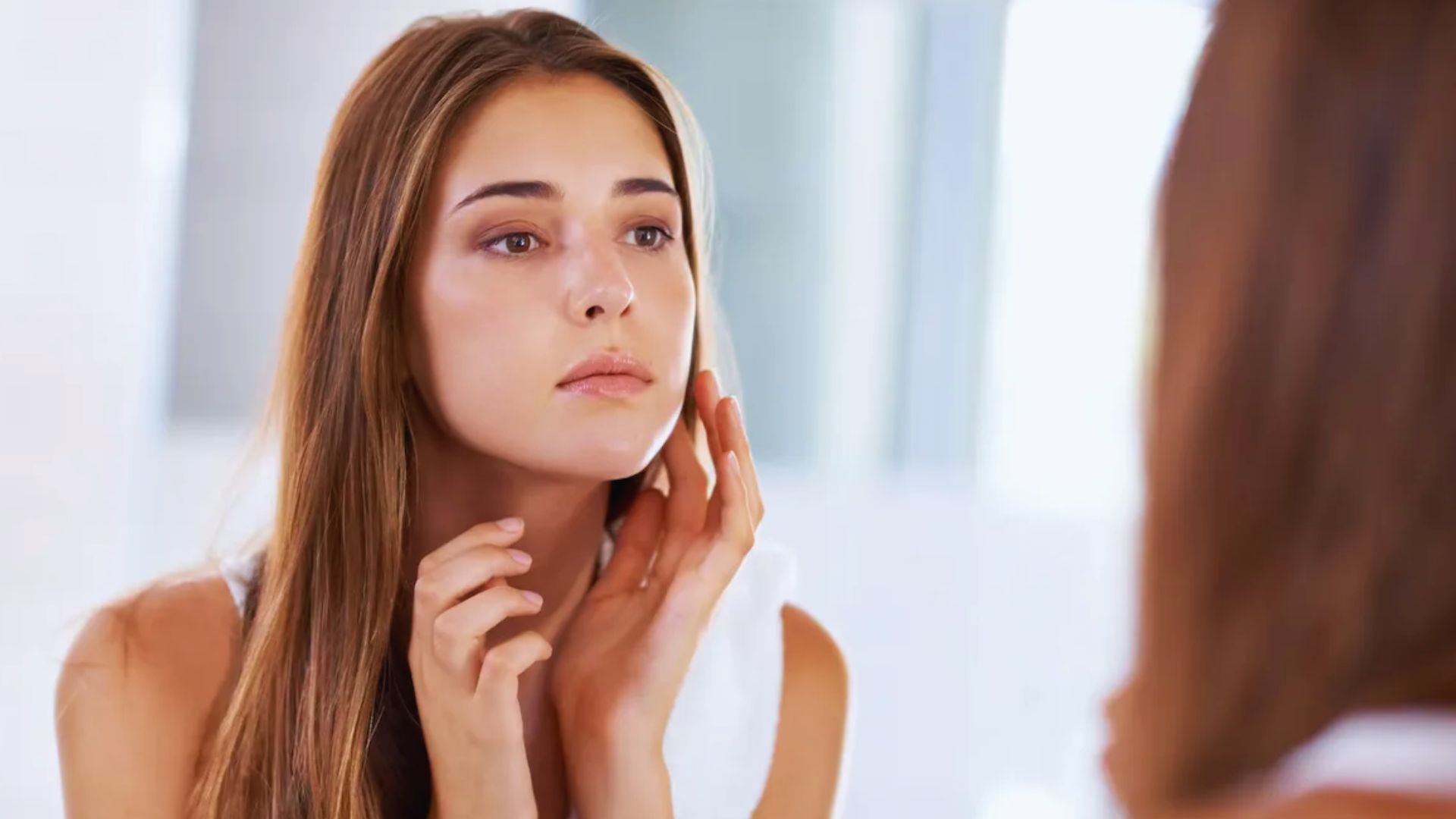 A woman with long hair examines her face in a mirror, gently touching her cheek. The setting is bright and serene, conveying a calm and introspective mood.