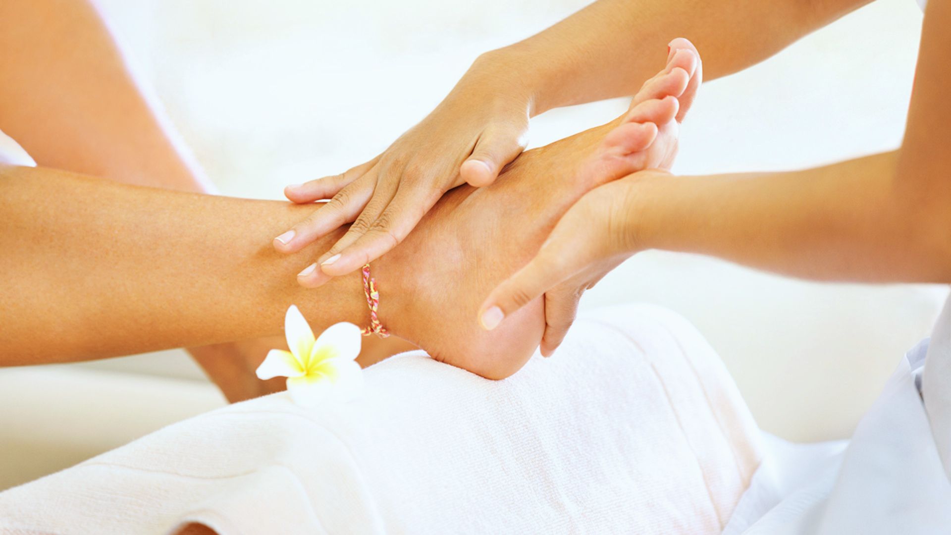 Close-up of a person receiving a relaxing foot massage. Hands gently press a foot adorned with an anklet and a white flower, conveying a serene spa atmosphere.