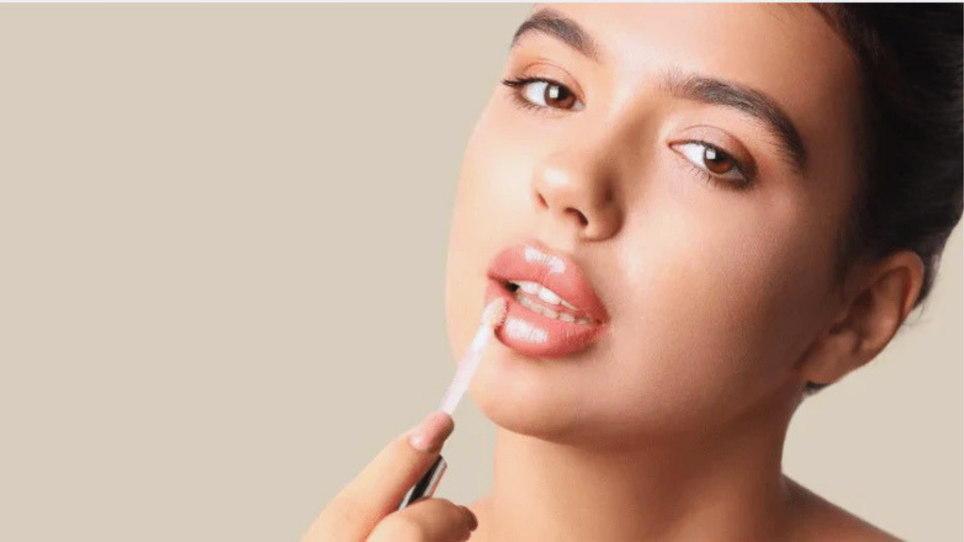 A person applies lip gloss with an applicator, set against a neutral background. The expression is calm, highlighting a natural makeup look.