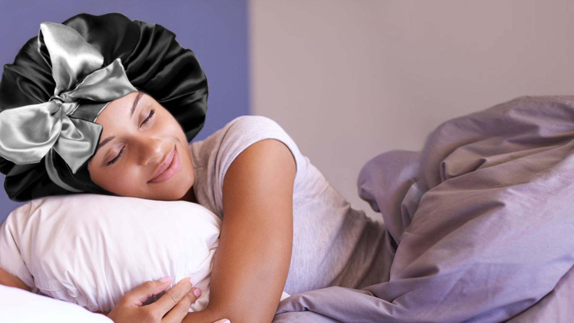 A person wearing a black satin bonnet with a silver bow smiles contentedly while asleep on a white pillow, wrapped in gray bedding. The scene conveys comfort and tranquility.