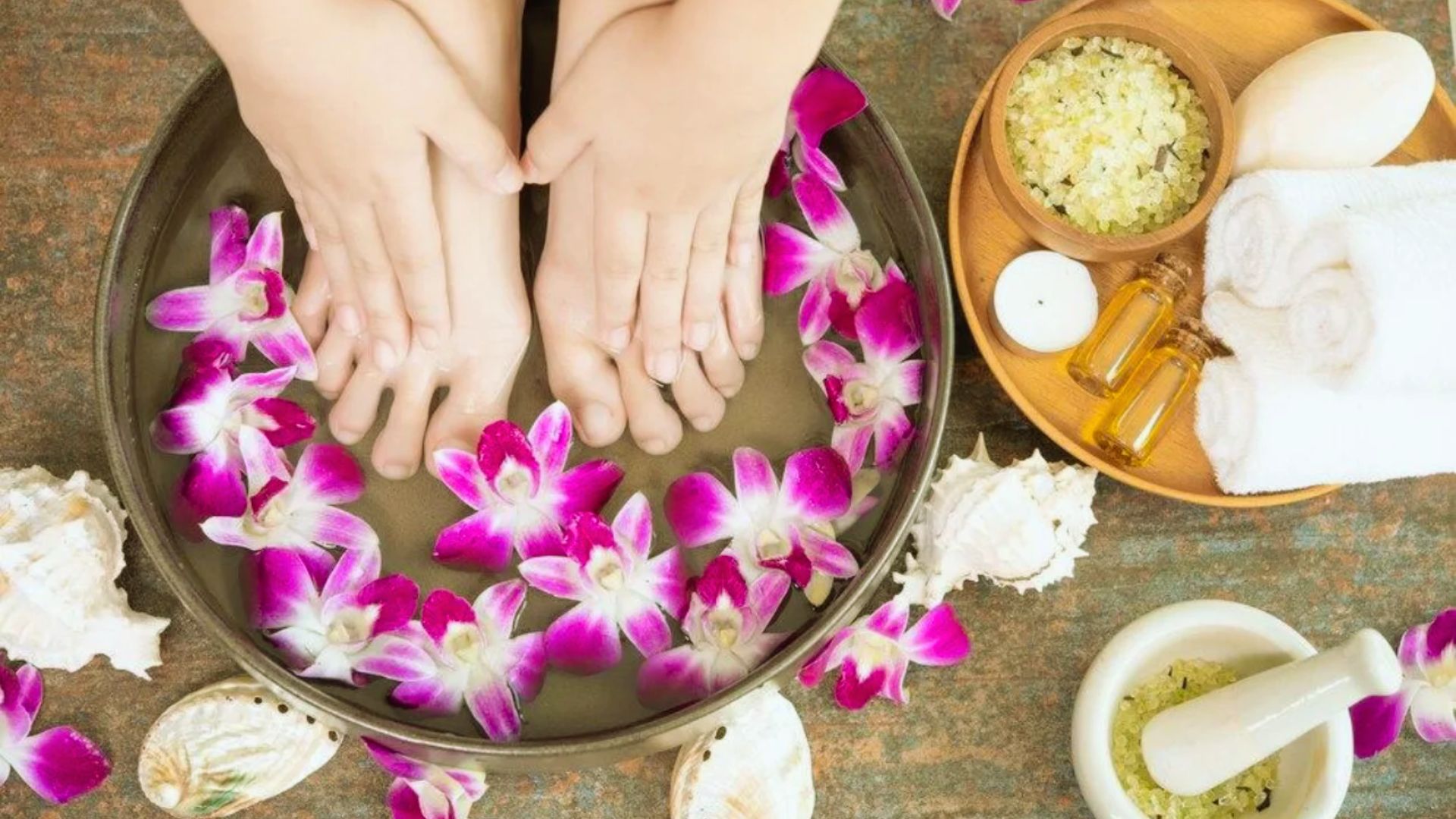 Feet soaking in a bowl filled with water and pink orchid petals. Nearby are sea shells, bath oils, a white towel, a candle, and a bowl of bath salts, creating a soothing spa ambiance.