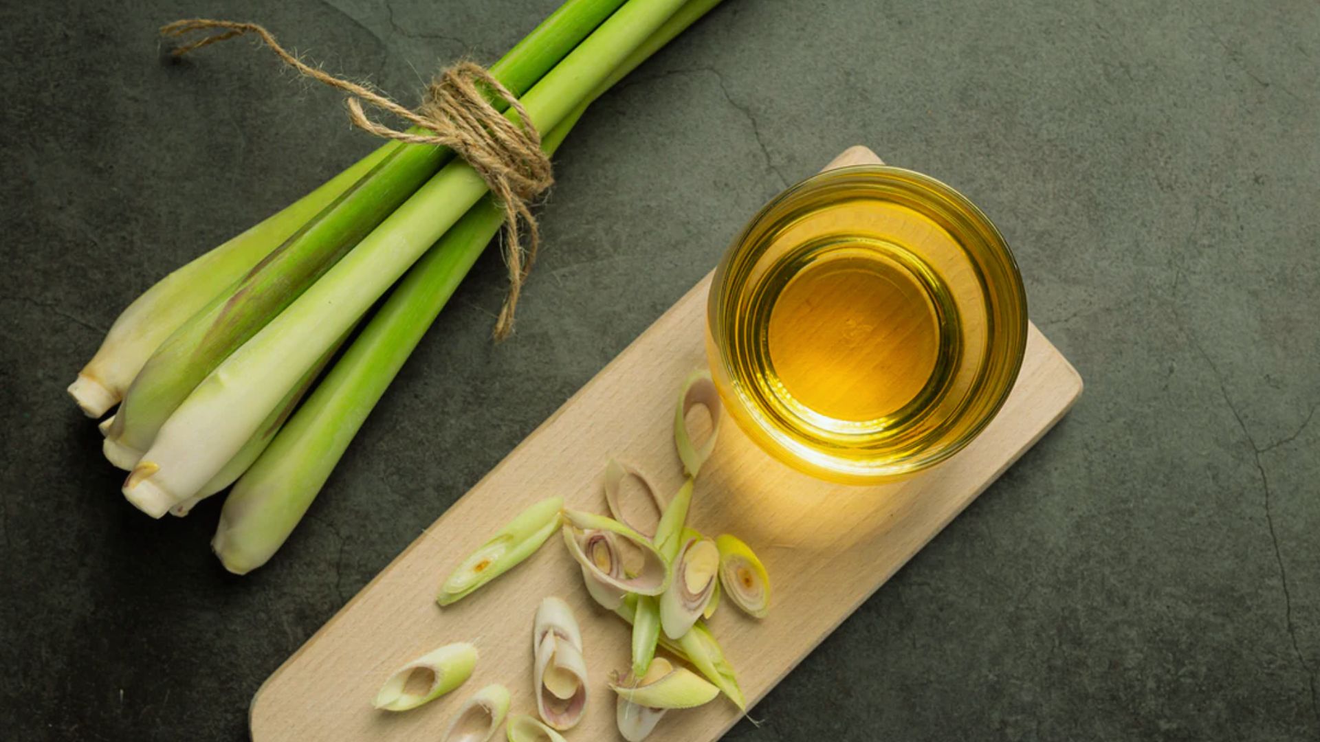 Fresh lemongrass stalks tied with twine, sliced lemongrass pieces, and a glass of golden oil on a wooden board. Dark, textured background.