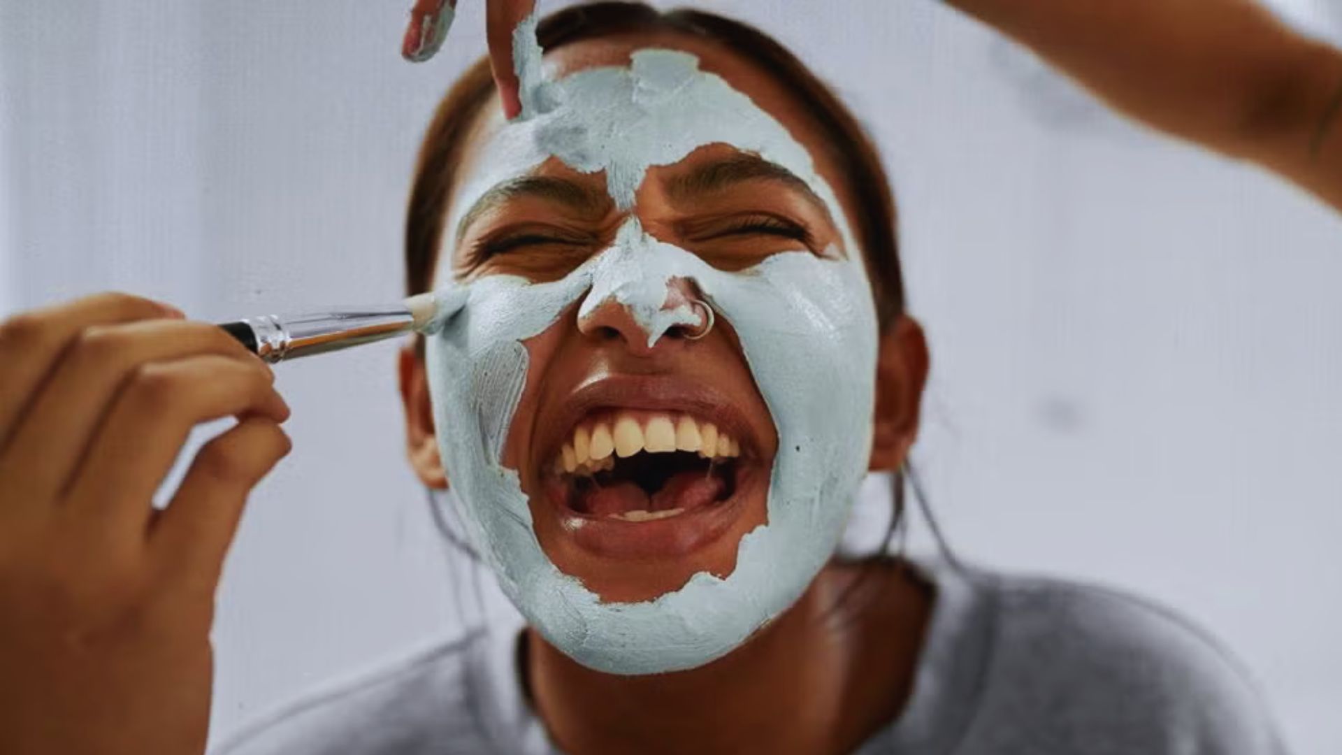 A person joyfully applies a blue face mask with a brush, laughing heartily. The scene conveys fun and relaxation in a skincare routine setting.