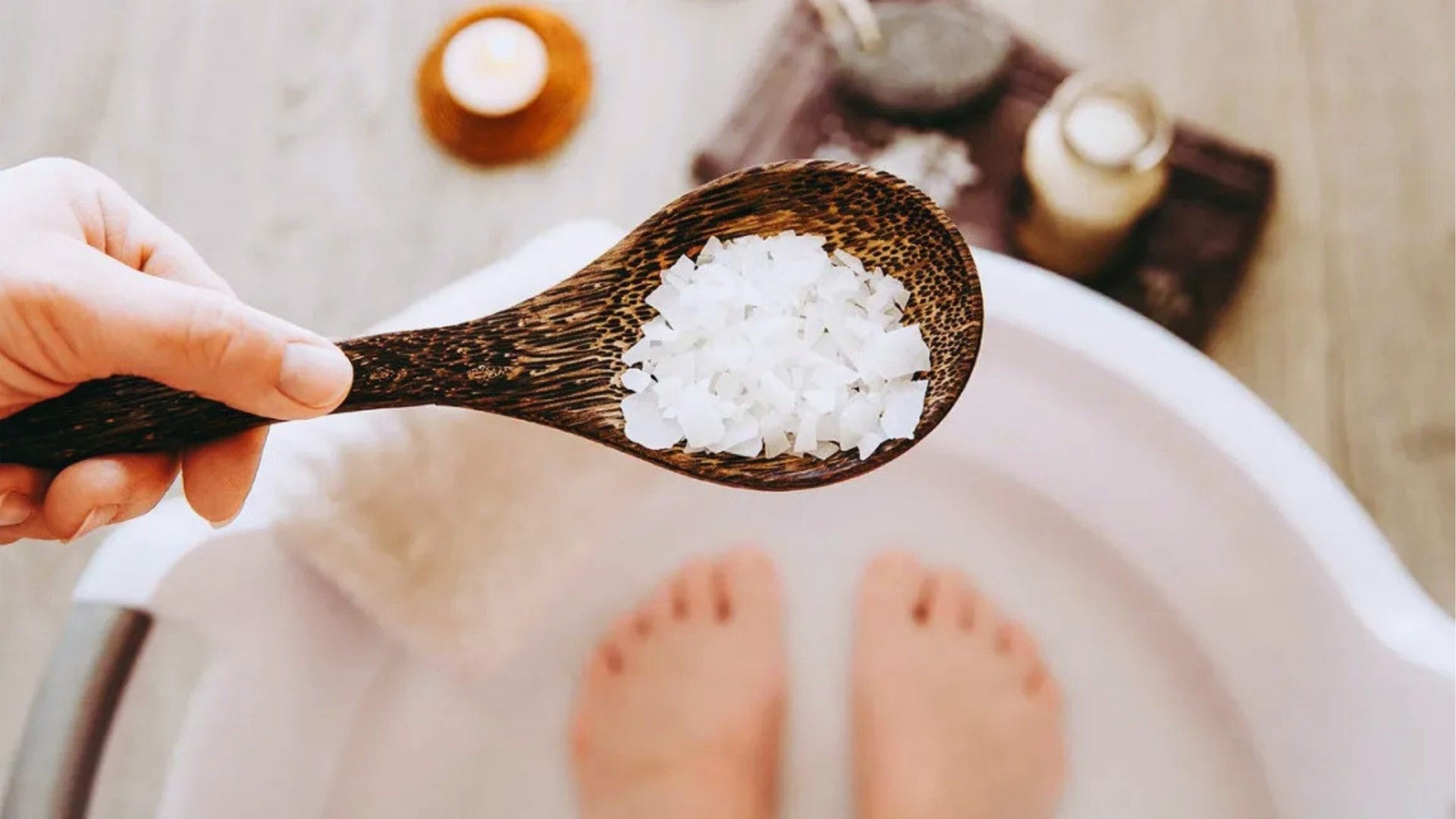 A hand holds a wooden spoon with bath salts above a foot soak. Blurred feet and spa items are visible in the background, creating a serene, relaxing ambiance.