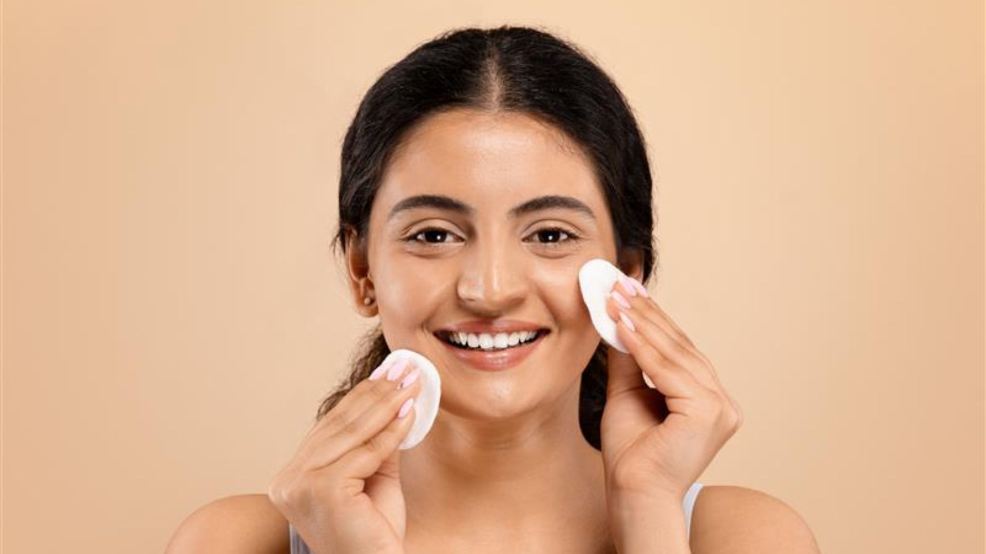 Smiling woman cleansing her face with cotton pads against a beige background. She appears relaxed and happy, engaging in a skincare routine.