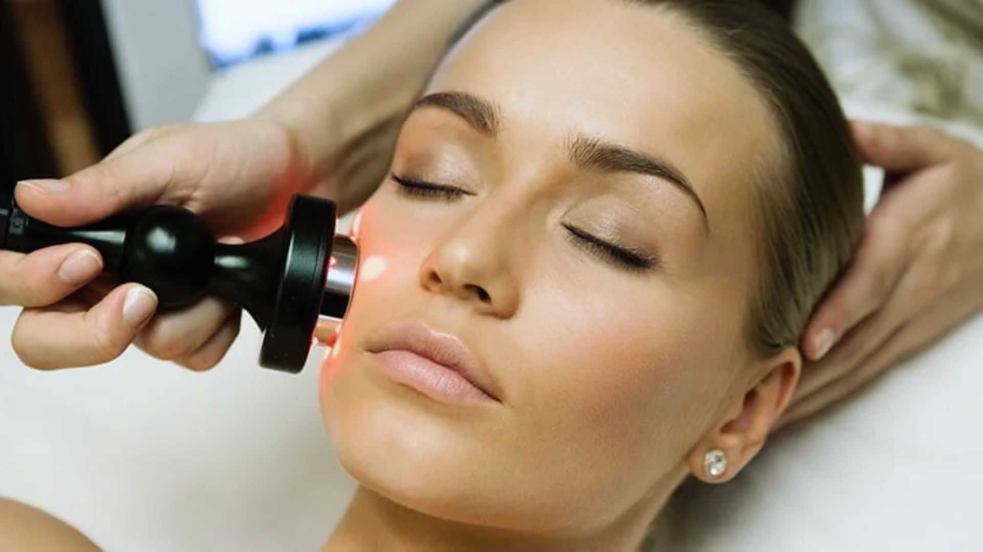 A woman receiving a skincare treatment; her eyes closed, calm expression, glowing red light on her cheek from a handheld device, creating a serene mood.