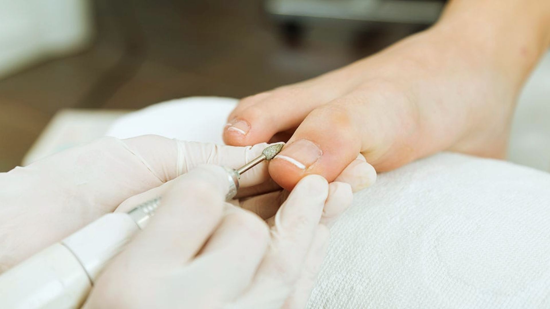 Close-up of a pedicure in progress. A gloved hand uses an electric file on a person's toenail, creating a relaxed and professional spa atmosphere.