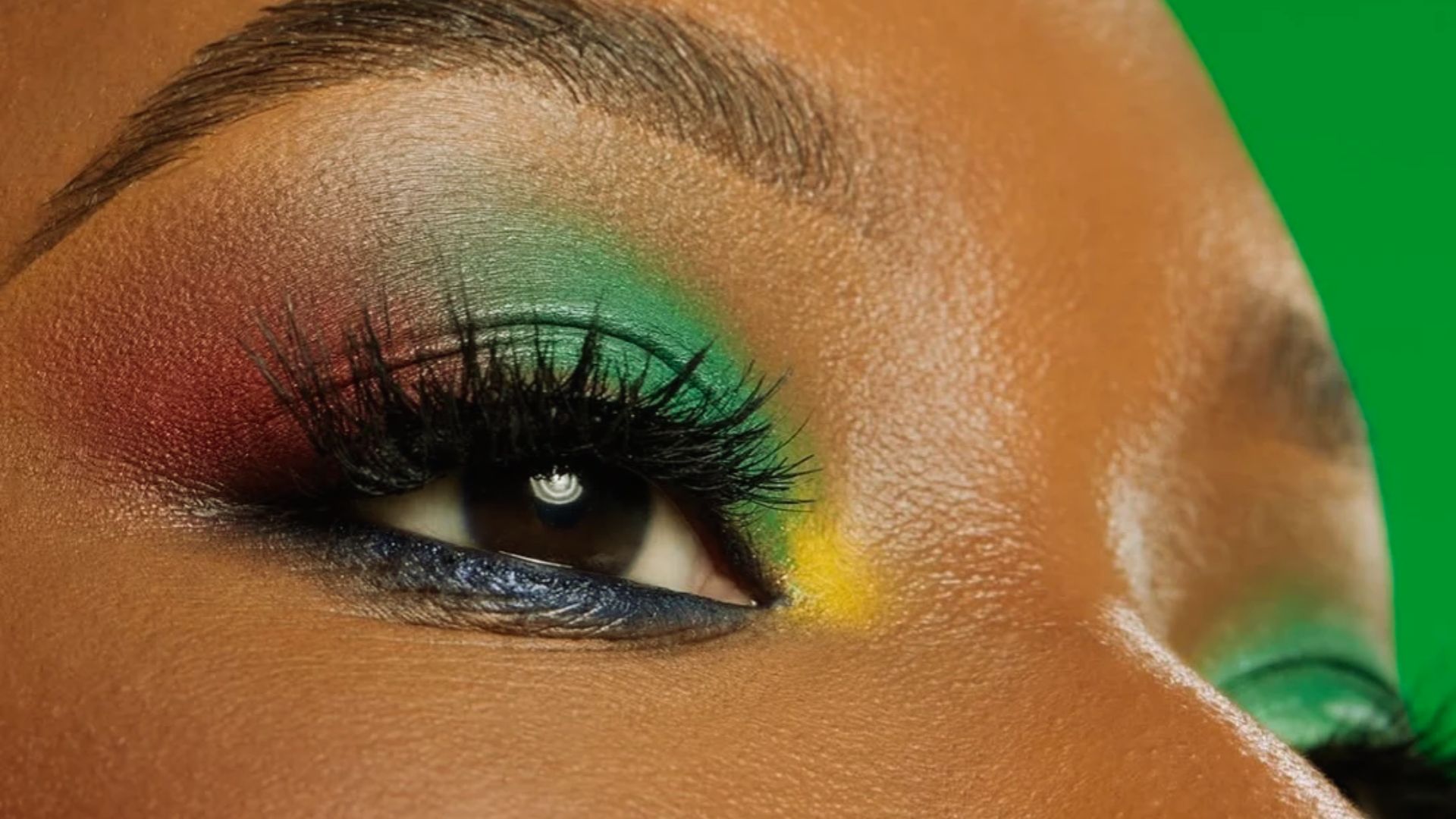 Close-up of a person's eye adorned with vibrant makeup. Features bold green, red, and yellow eyeshadow, long eyelashes, and a bright green background.