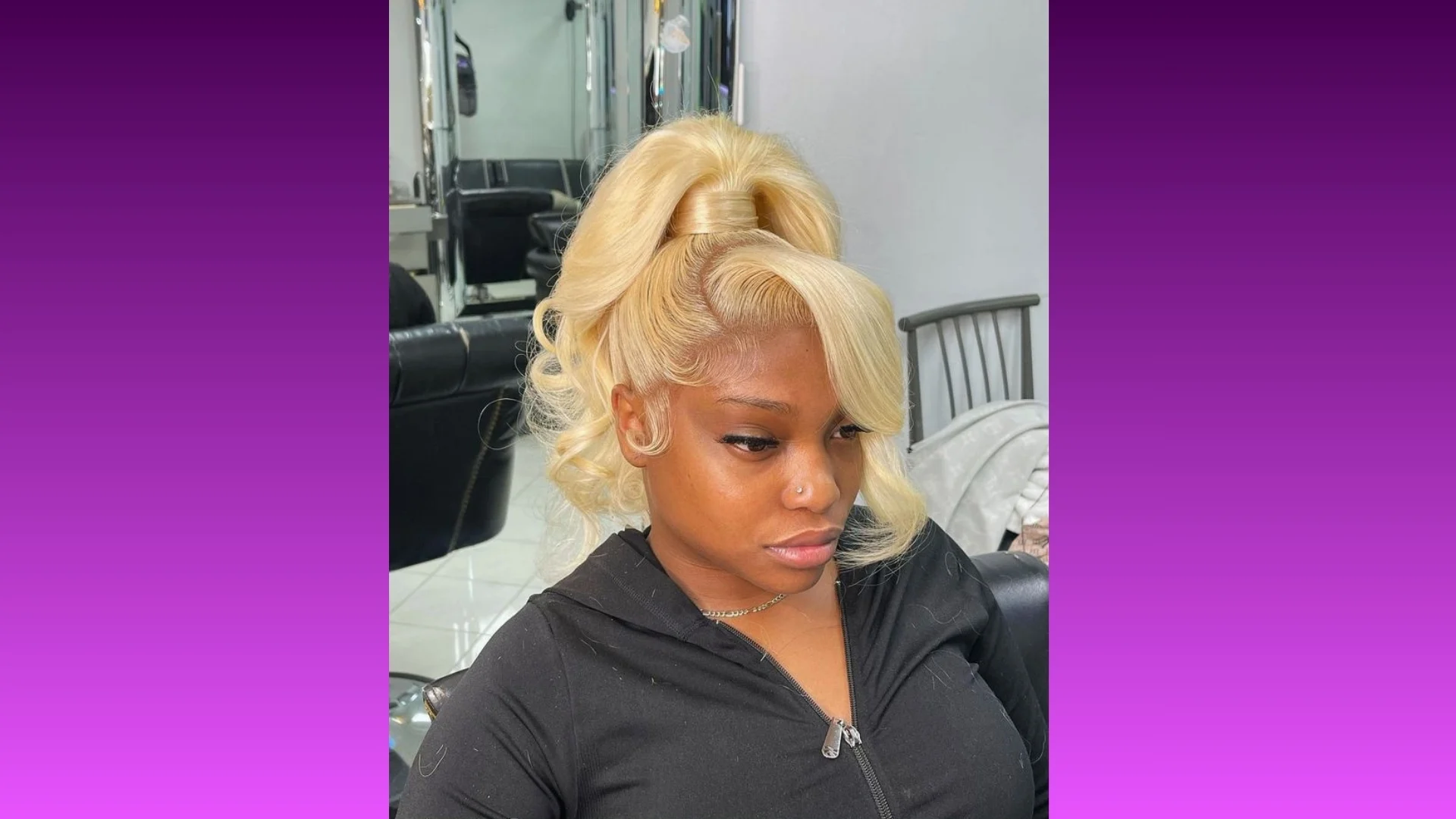 A woman with a high blonde ponytail and curled tendrils sits in a salon, wearing a black zip-up hoodie. The background is a gradient of purple shades.