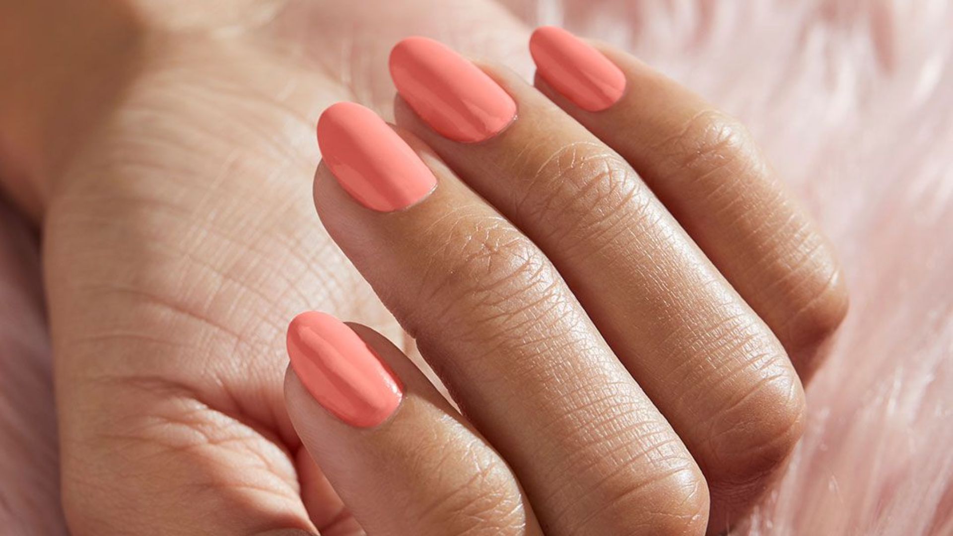 Close-up of a hand with glossy peach-colored nail polish on neatly manicured nails, set against a soft, pink, textured background, conveying elegance.