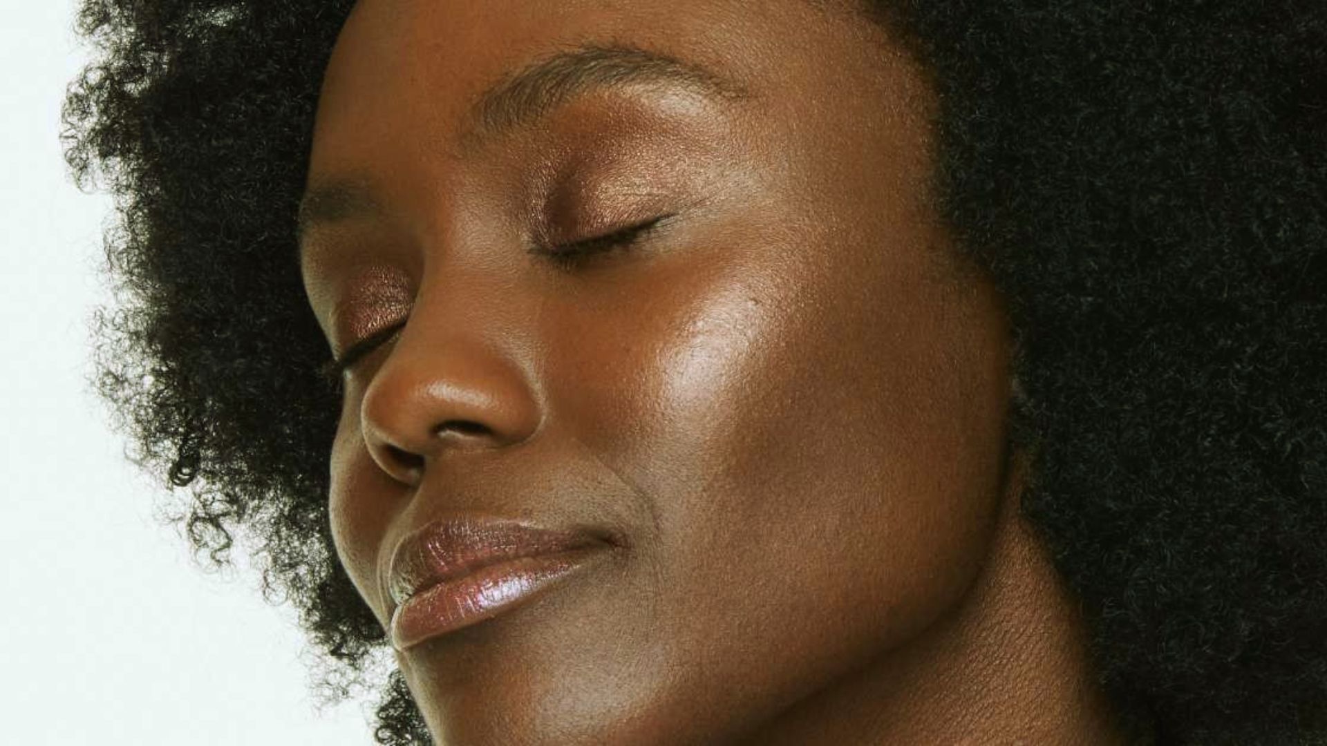 A woman with closed eyes and an expression of calm. She has natural, curly hair and glowing skin, conveying a serene and peaceful mood.