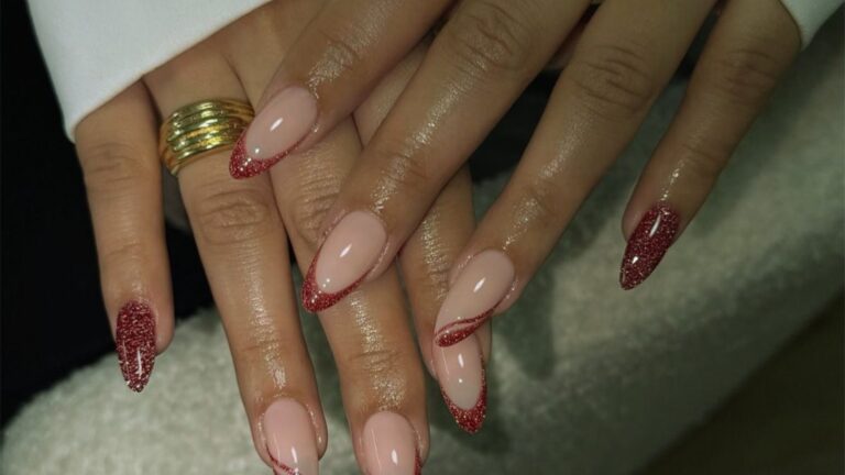 Close-up of hands with almond-shaped nails, featuring nude polish and red glitter accents. The right hand wears a gold ring. The tone is elegant and stylish.