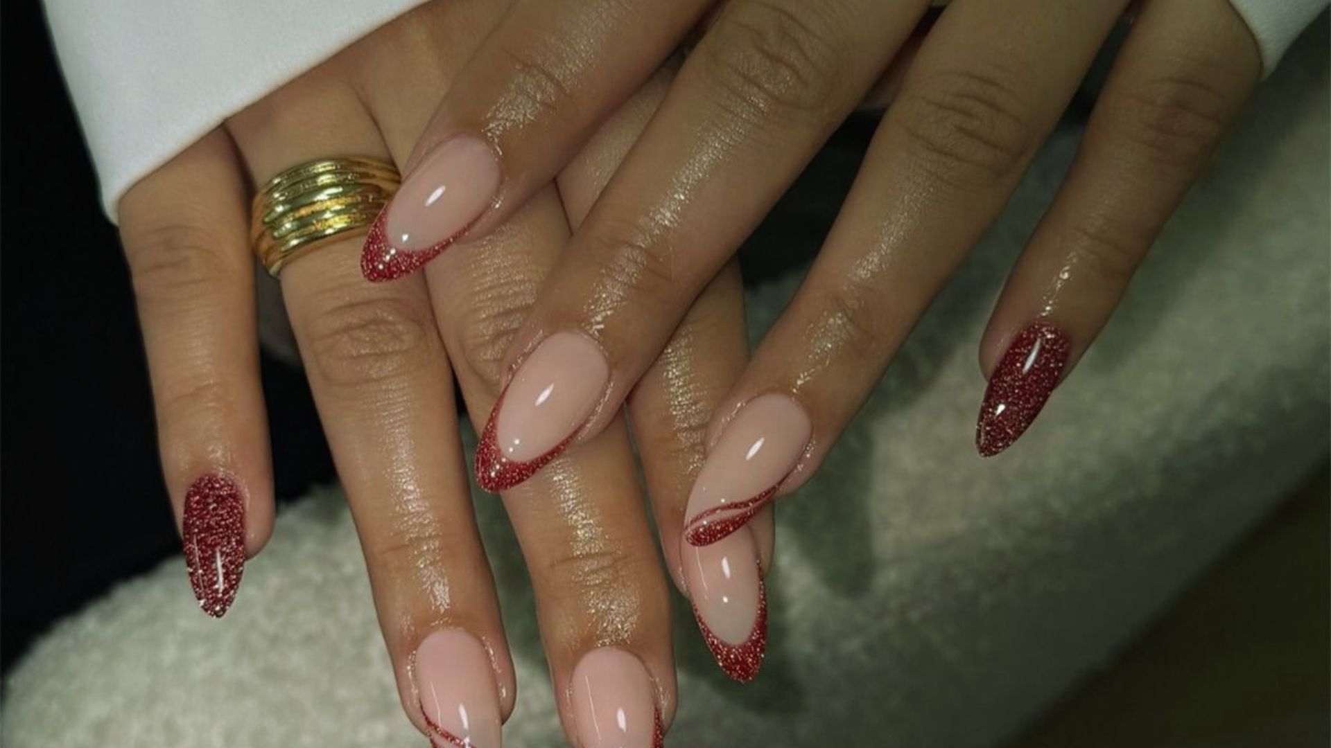 Close-up of hands with almond-shaped nails, featuring nude polish and red glitter accents. The right hand wears a gold ring. The tone is elegant and stylish.