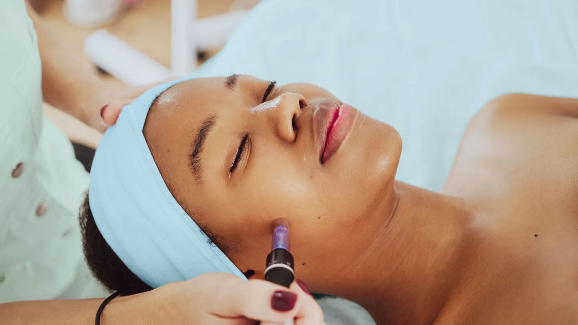 A woman enjoys a relaxing facial treatment, eyes closed, with a blue headband. Aesthetician uses a small device on her cheek. Peaceful ambiance.