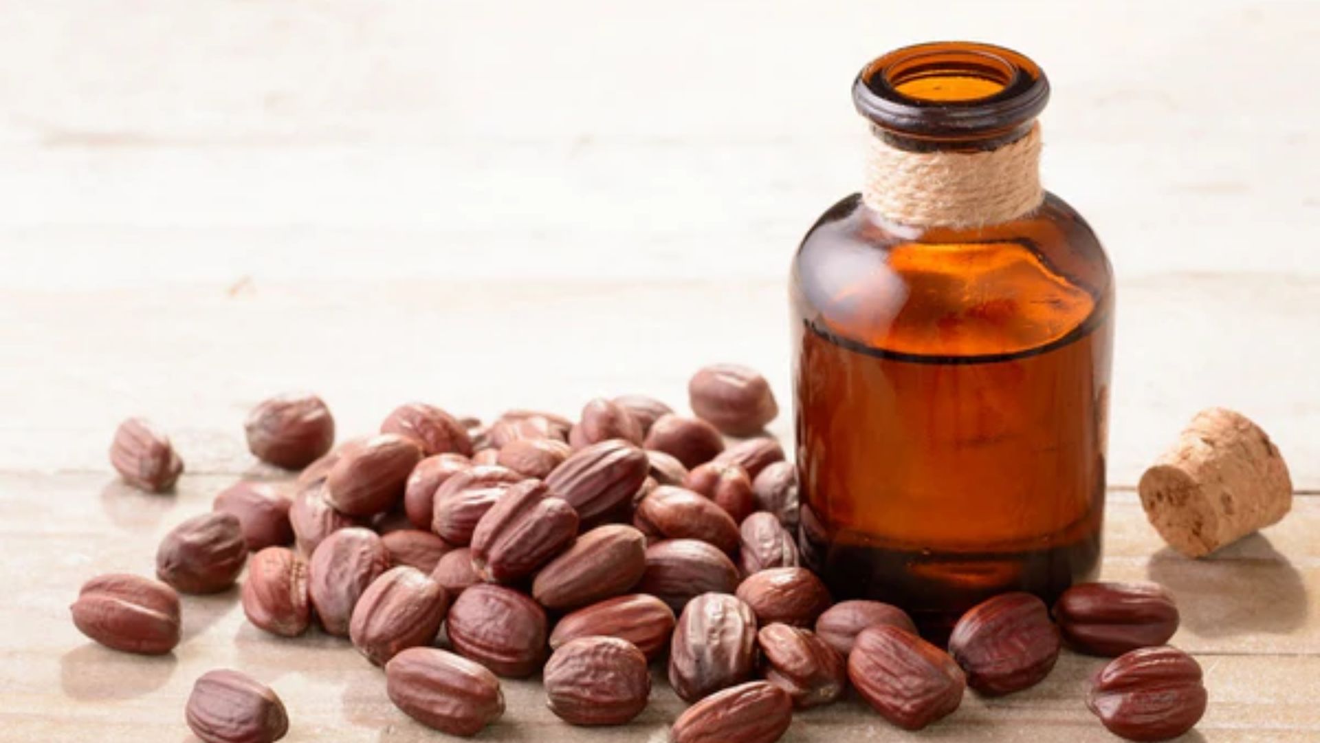 A small amber glass bottle of oil sits on a light wooden surface, surrounded by scattered brown jojoba seeds. The cork lies next to the bottle. Rustic and natural.