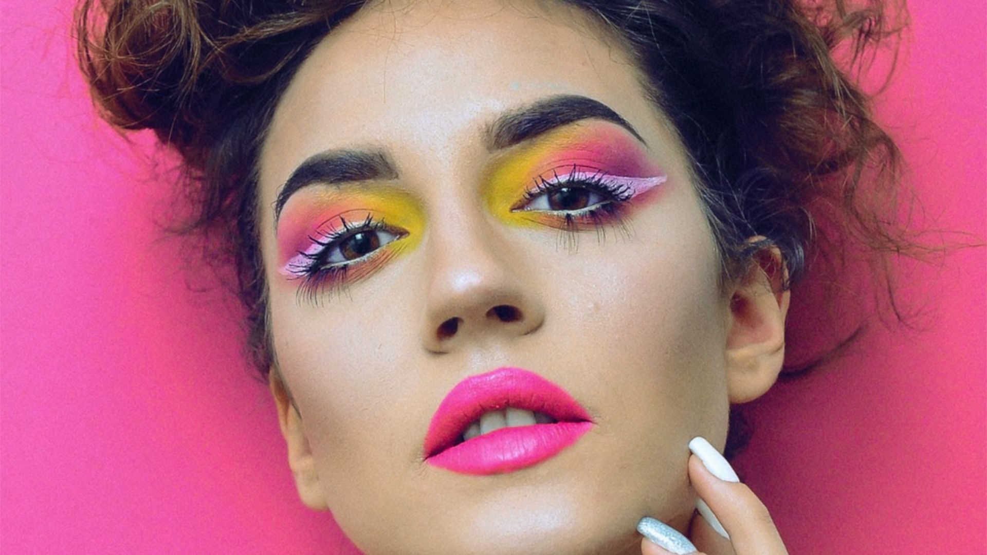 Woman with vibrant makeup against a pink background. Her eyeshadow blends yellow and pink, with bold pink lips and styled curly hair, creating a striking, artistic look.
