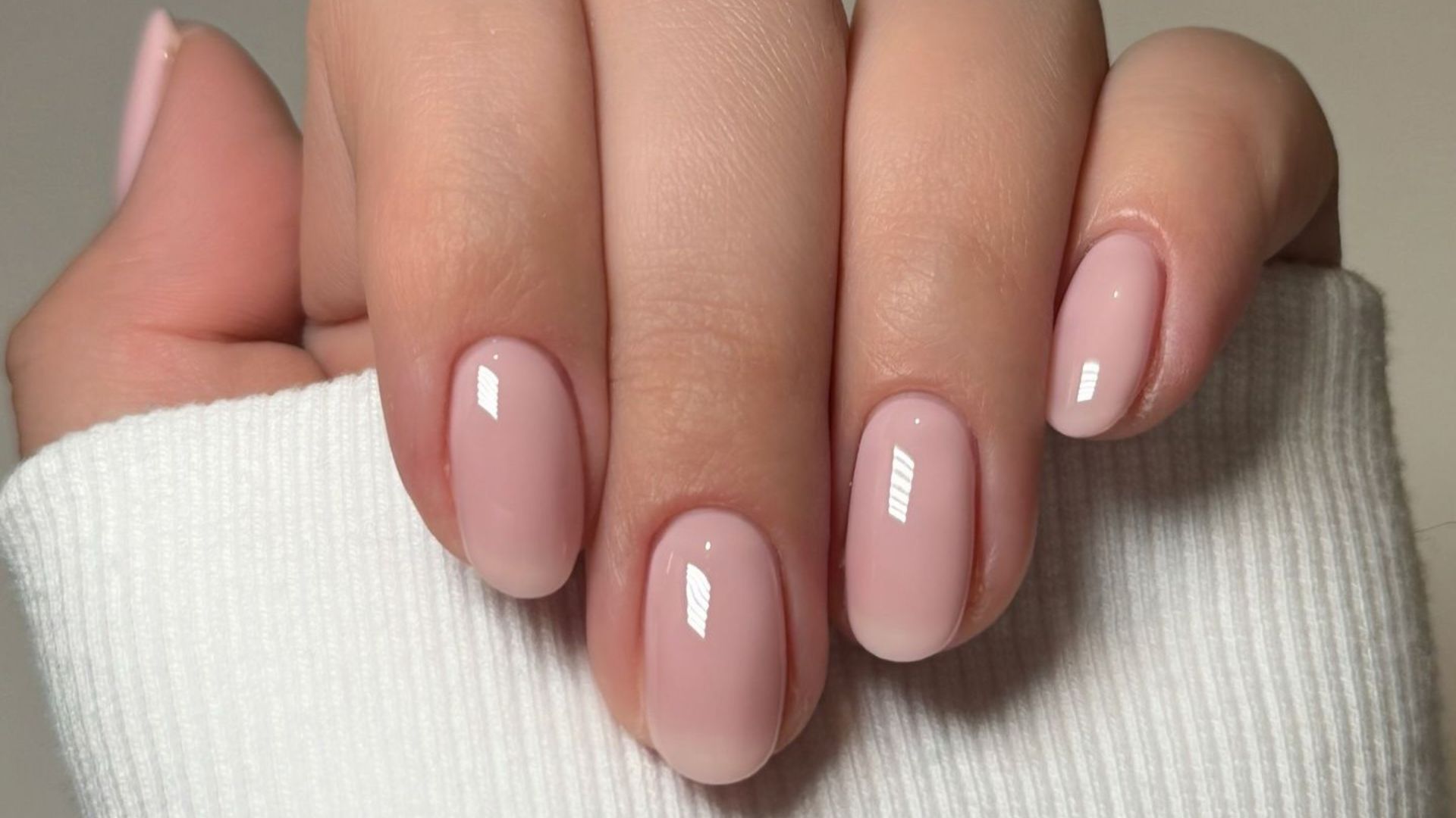 Close-up of a hand with neatly manicured nails painted in a glossy, soft pink shade. The hand is partially covered by a white, ribbed fabric, giving a cozy feel.