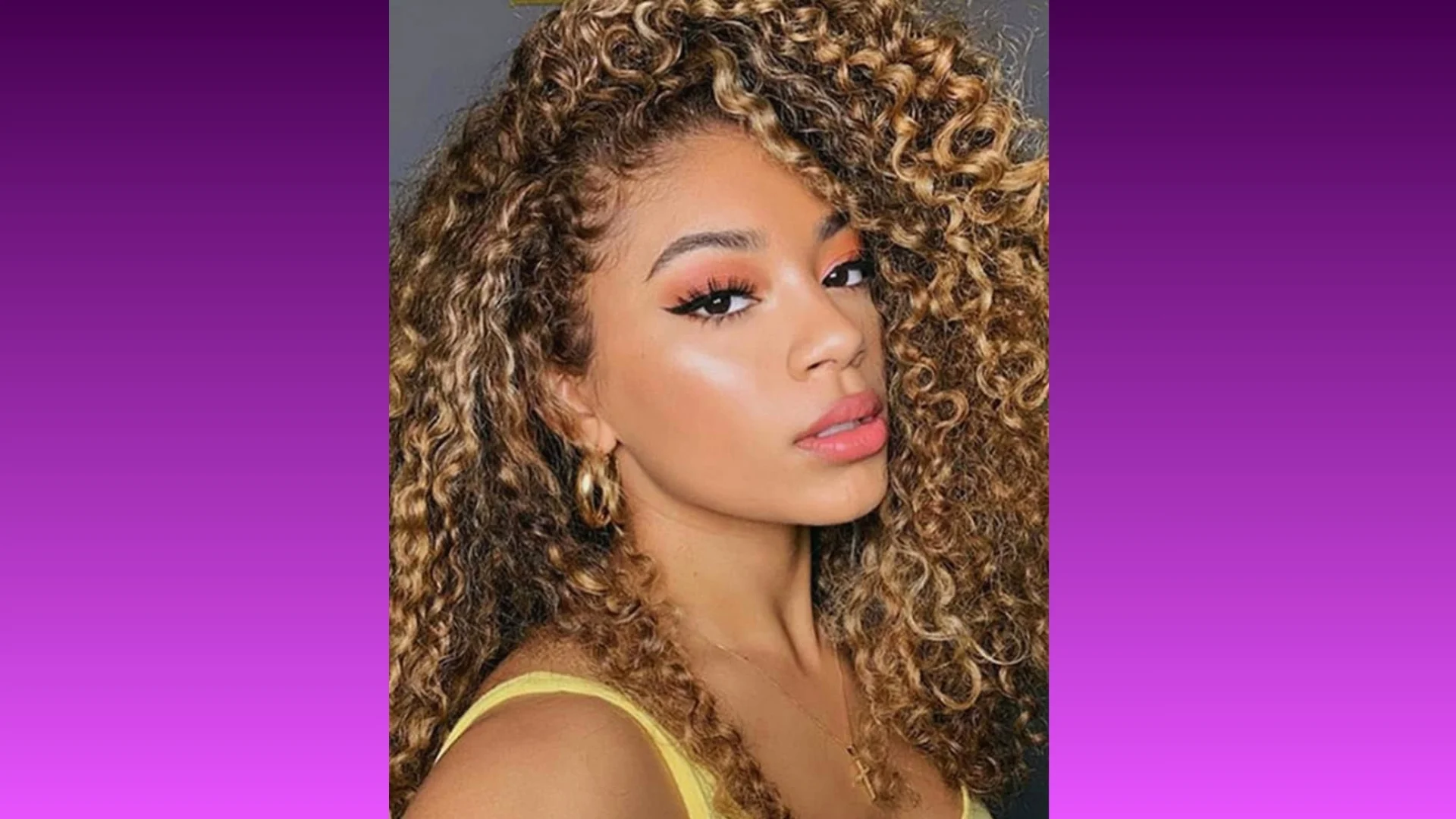 Portrait of a person with voluminous curly hair and vibrant makeup, wearing a yellow top and hoop earrings, set against a gradient purple background.