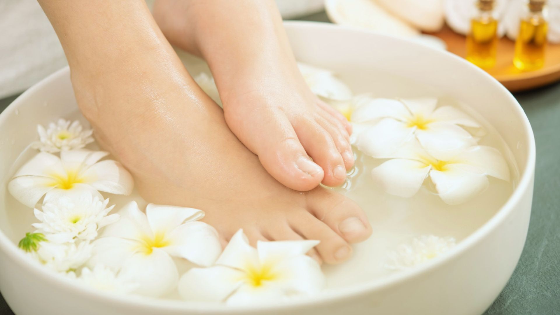 Feet soaking in a bowl of water with floating white flowers, creating a serene spa-like atmosphere. Nearby, small bottles of essential oils suggest relaxation.