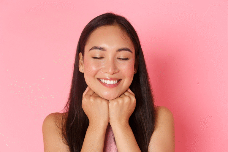 Beautiful Asian woman smiling with her eyes closed against a soft pink background.