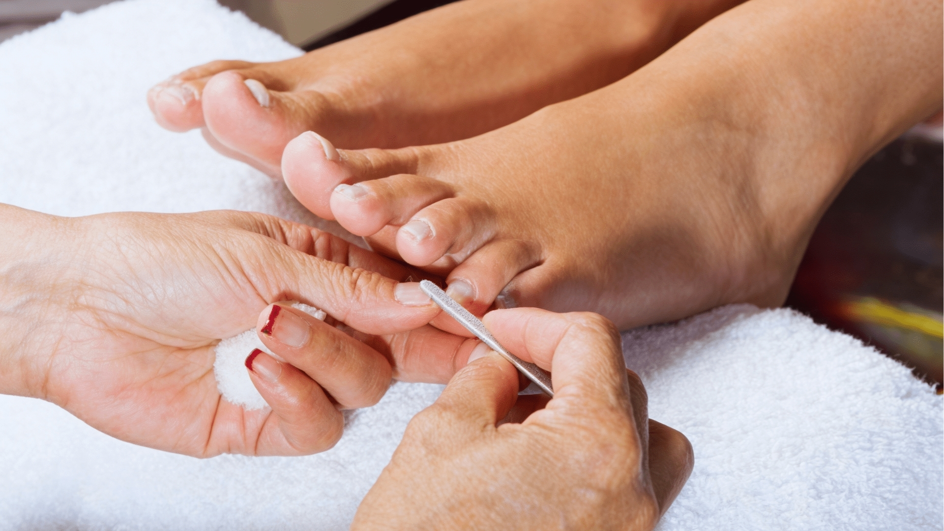 Close-up of a pedicure in progress. A pair of hands is carefully shaping a toenail with a metal file, set against a soft white towel. Relaxing atmosphere.