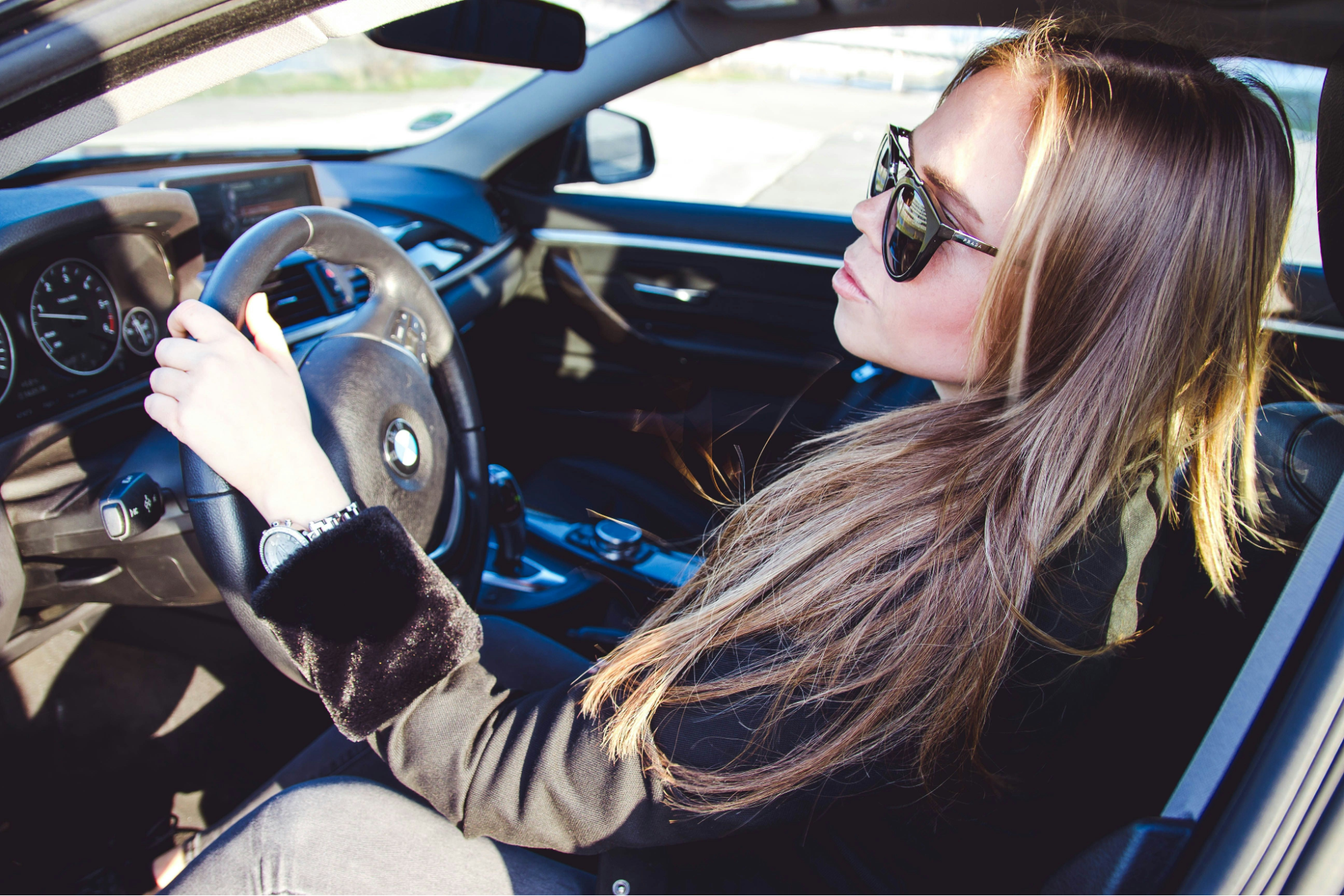 A woman with long hair wearing sunglasses is sitting confidently in a car, holding the steering wheel. The interior is sleek and modern.