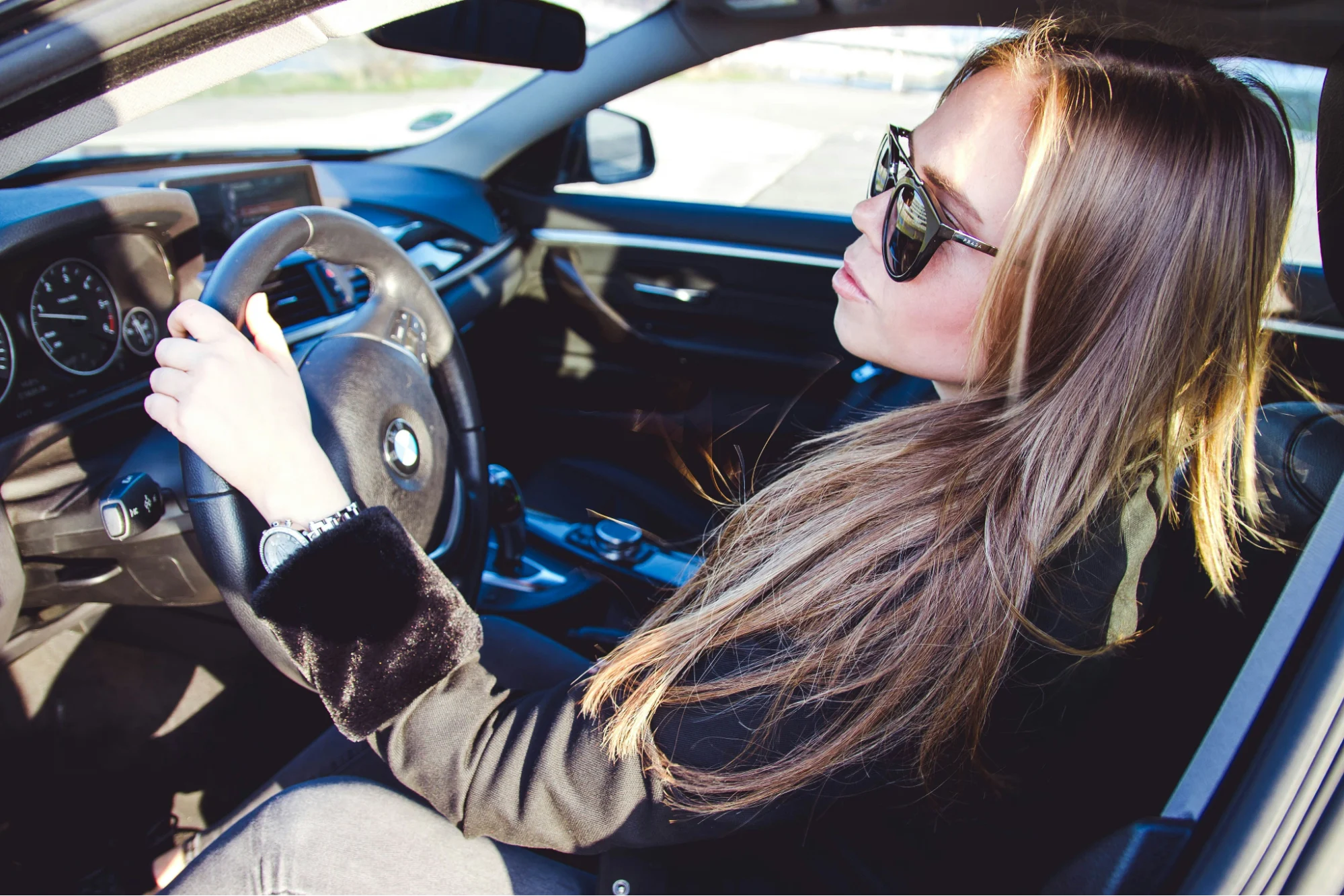 A woman with long hair wearing sunglasses is sitting confidently in a car, holding the steering wheel. The interior is sleek and modern.