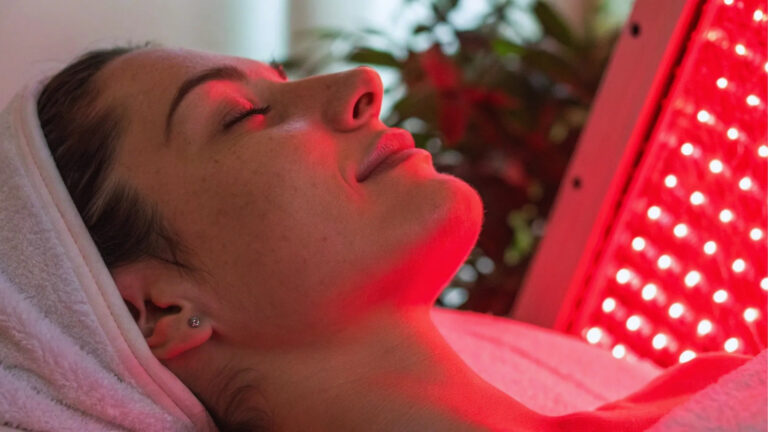 A person relaxes with closed eyes under a red LED light therapy panel, wrapped in a white towel. The scene conveys a calm, serene atmosphere.