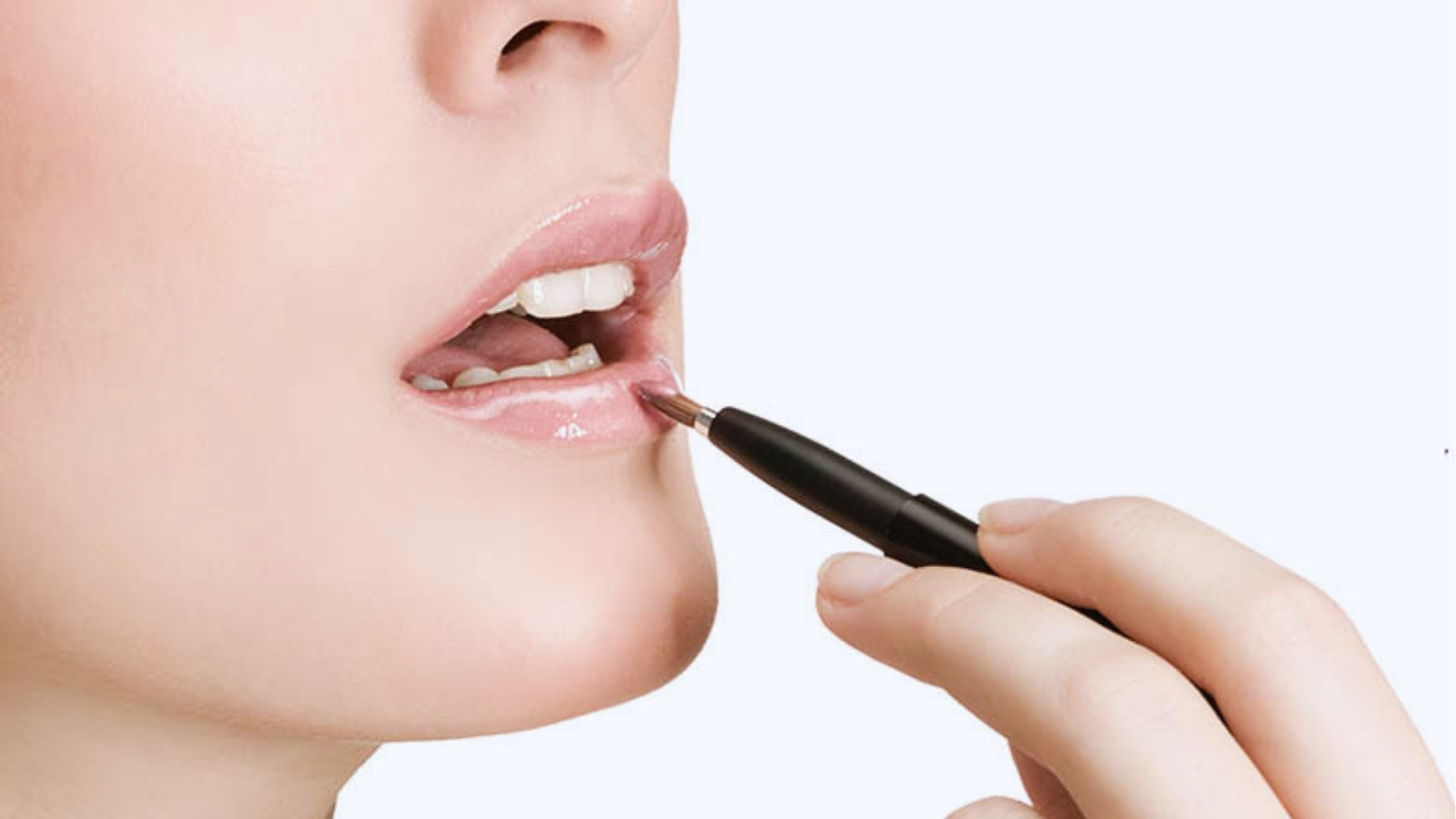 A person applies glossy lipstick with a brush. The close-up shows their lips slightly parted, conveying a sense of care and precision in makeup application.