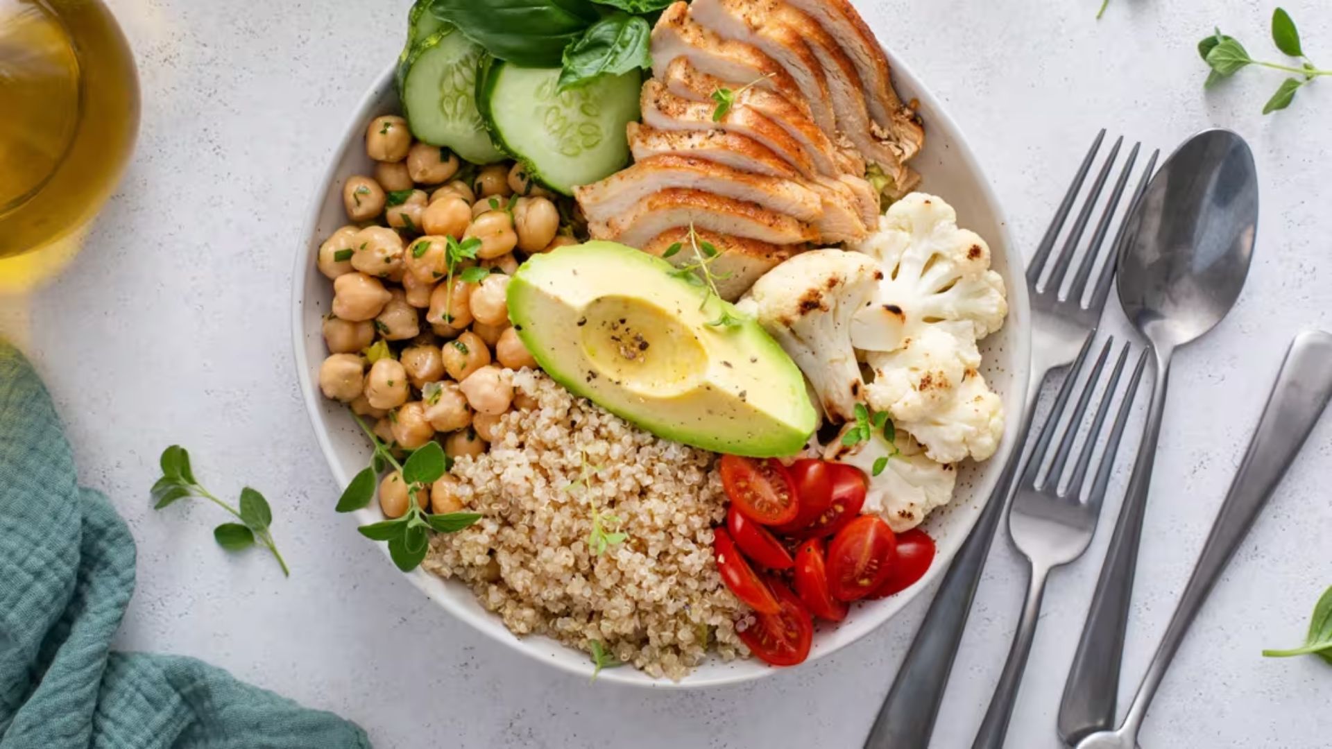 A vibrant salad bowl with sliced grilled chicken, quinoa, avocado, chickpeas, cucumbers, cherry tomatoes, and cauliflower, garnished with herbs on a light background.