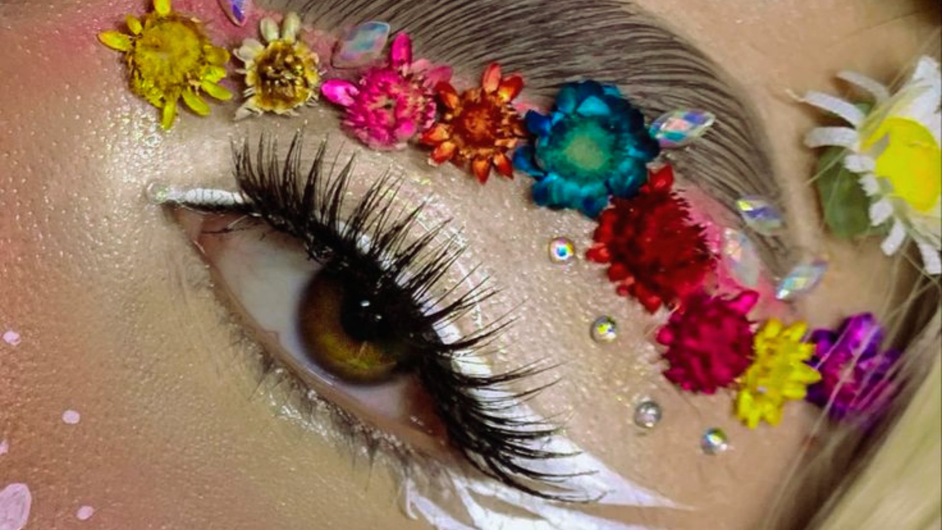 Close-up of an eye with vibrant makeup. Dried colorful flowers and sparkly gems adorn the eyebrow area, emphasizing long lashes and white eyeliner. Playful and artistic mood.
