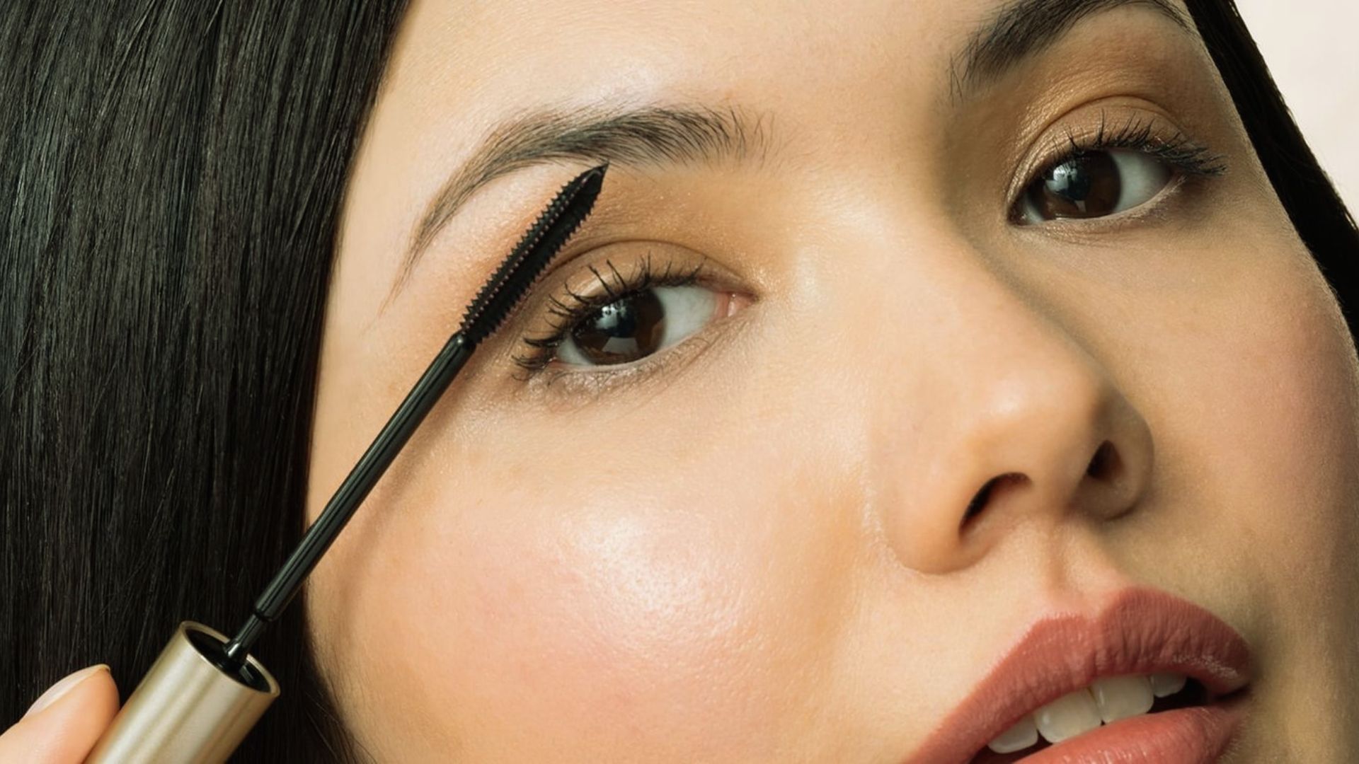 Close-up of a woman applying mascara with a gold brush. Her skin is glowing, lips slightly parted, and eyes focused, conveying a calm and confident mood.