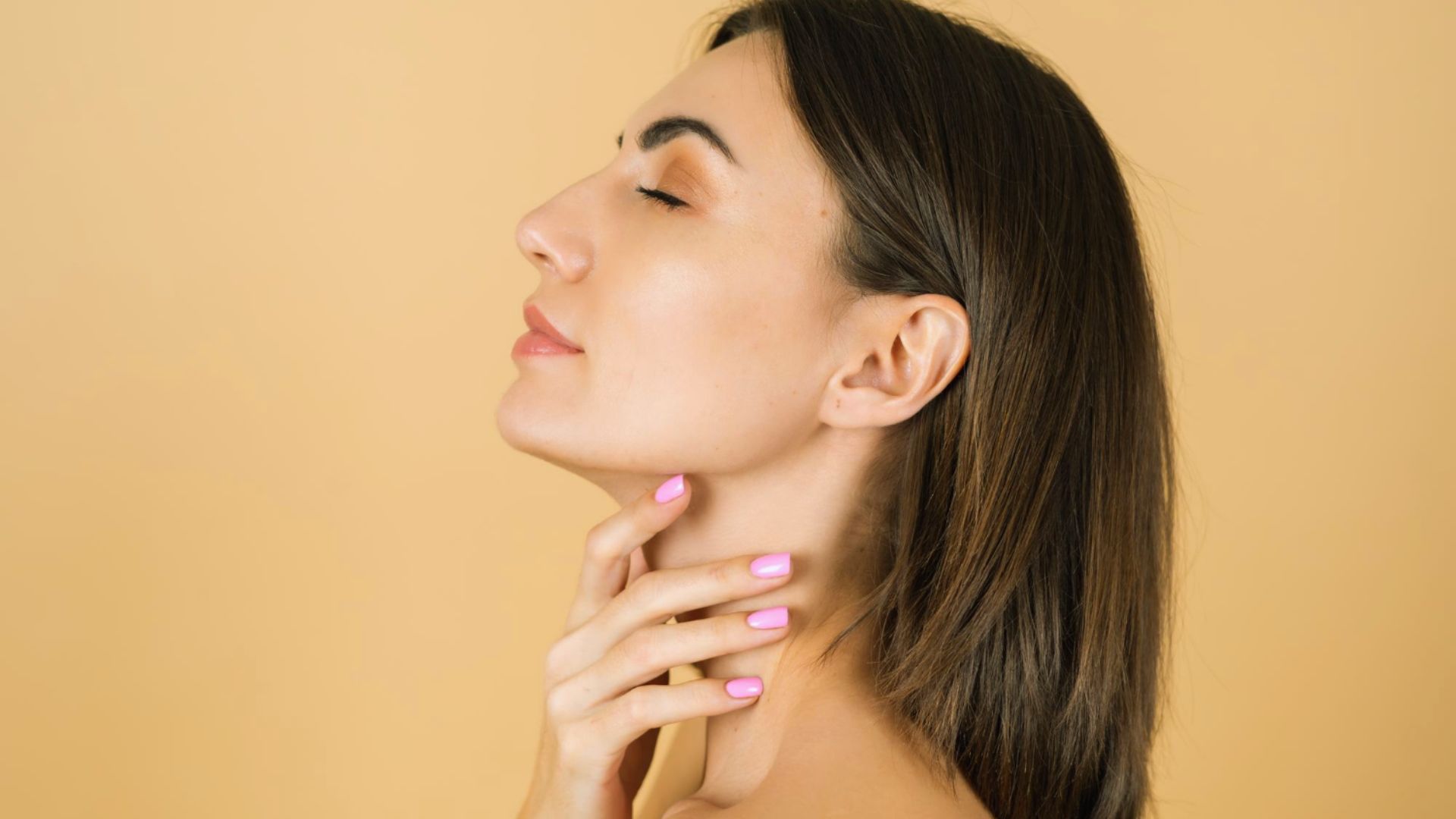 Side profile of a woman with long brown hair against a soft beige background. She gently touches her neck, eyes closed, exuding a serene and relaxed mood.