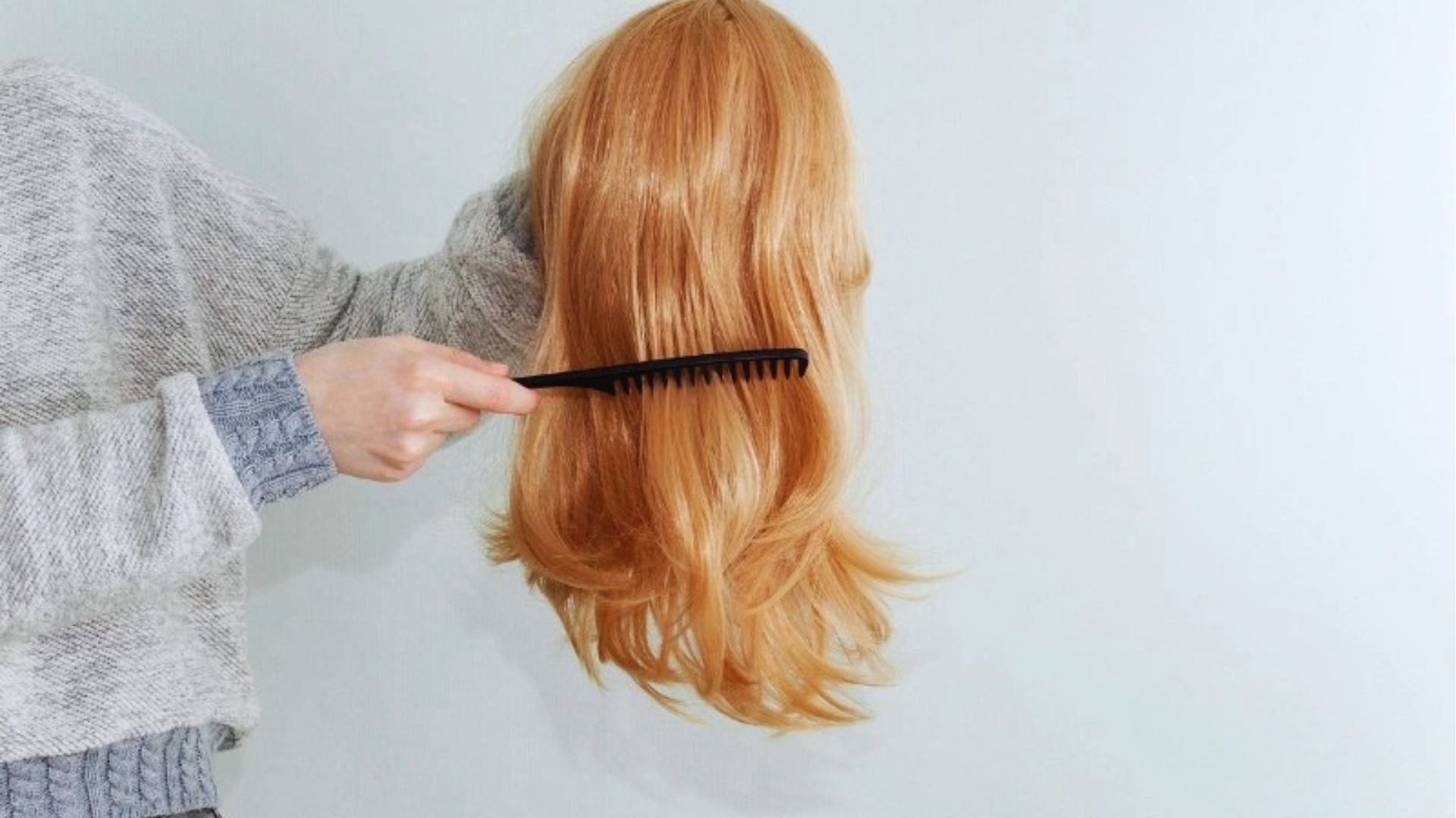 A person in a gray sweater holds a blonde wig, gently combing it with a black comb. The background is a light, solid color, creating a calm atmosphere.