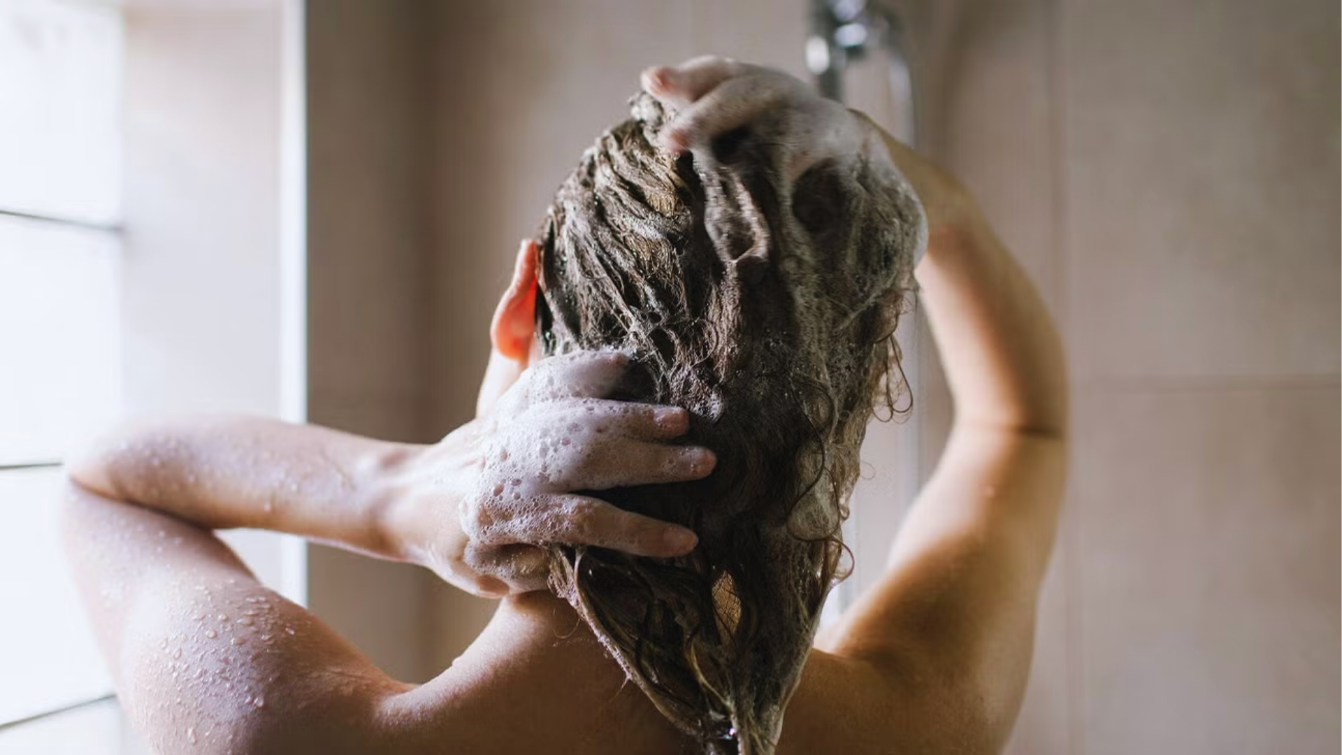 Person washing their hair in the shower, covered in soap suds. Hands massage the scalp, water droplets visible on skin, conveying a sense of relaxation.
