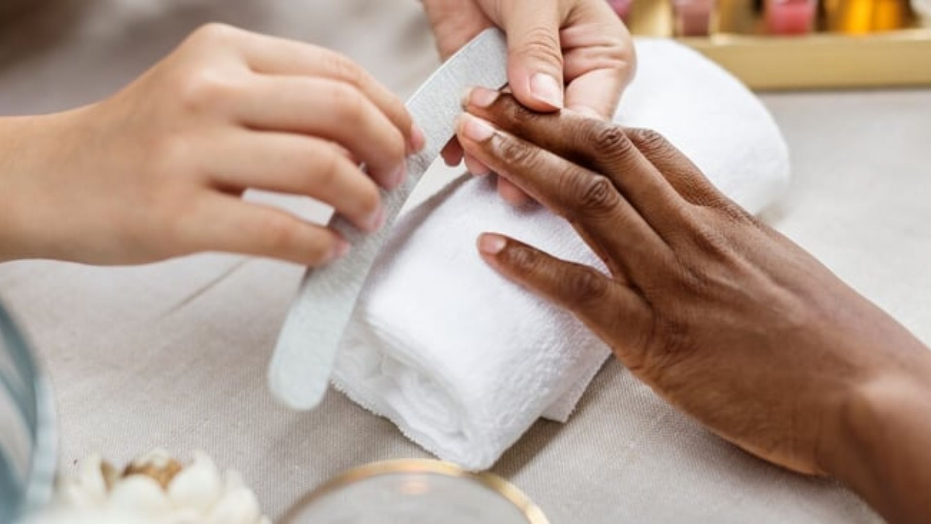 A person receives a manicure, with one hand being filed while resting on a white towel. The scene conveys a calm, spa-like atmosphere.